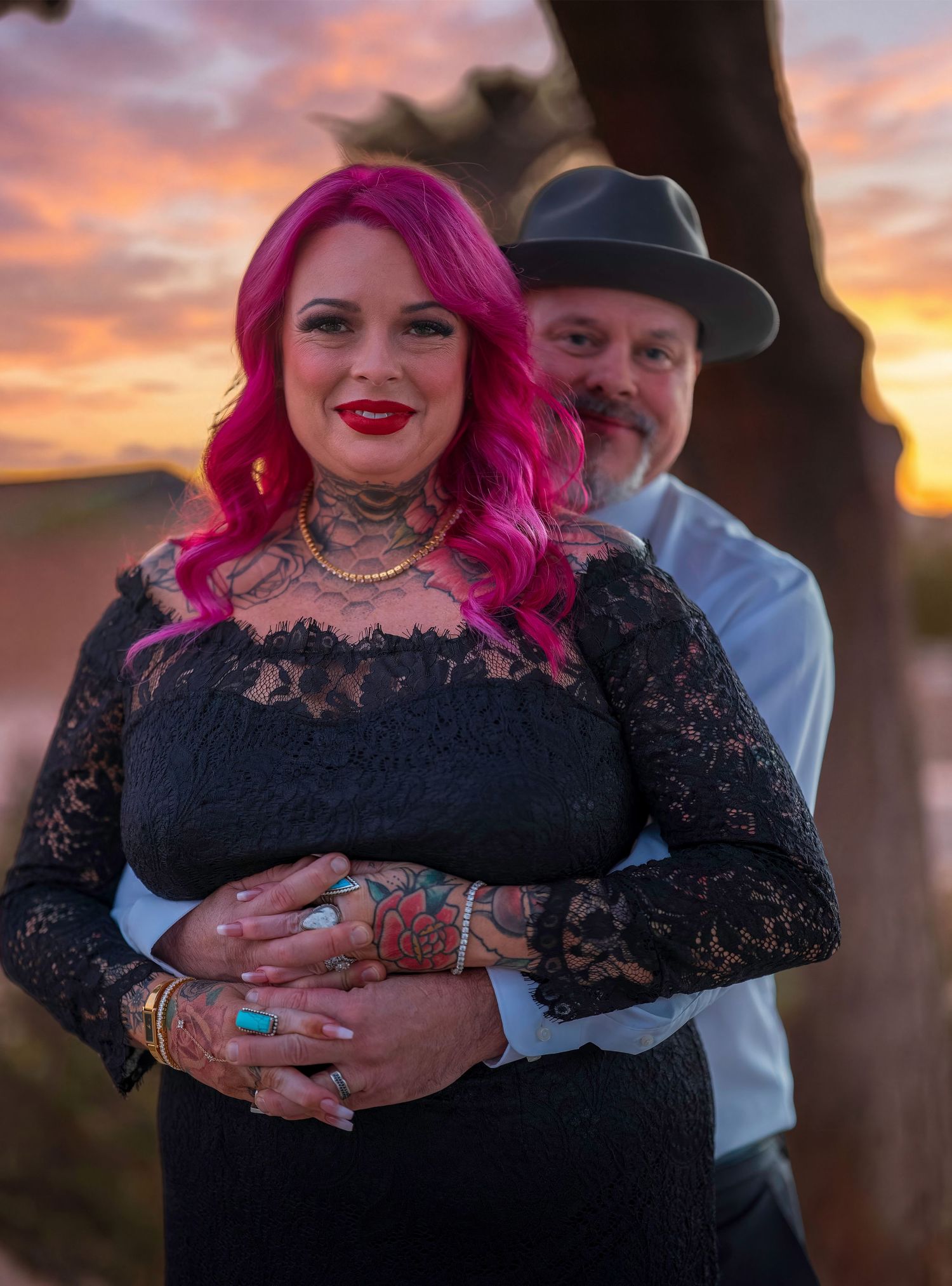 Couple posing at sunset, woman with pink hair and tattoos in black lace dress, man in gray hat behind her.
