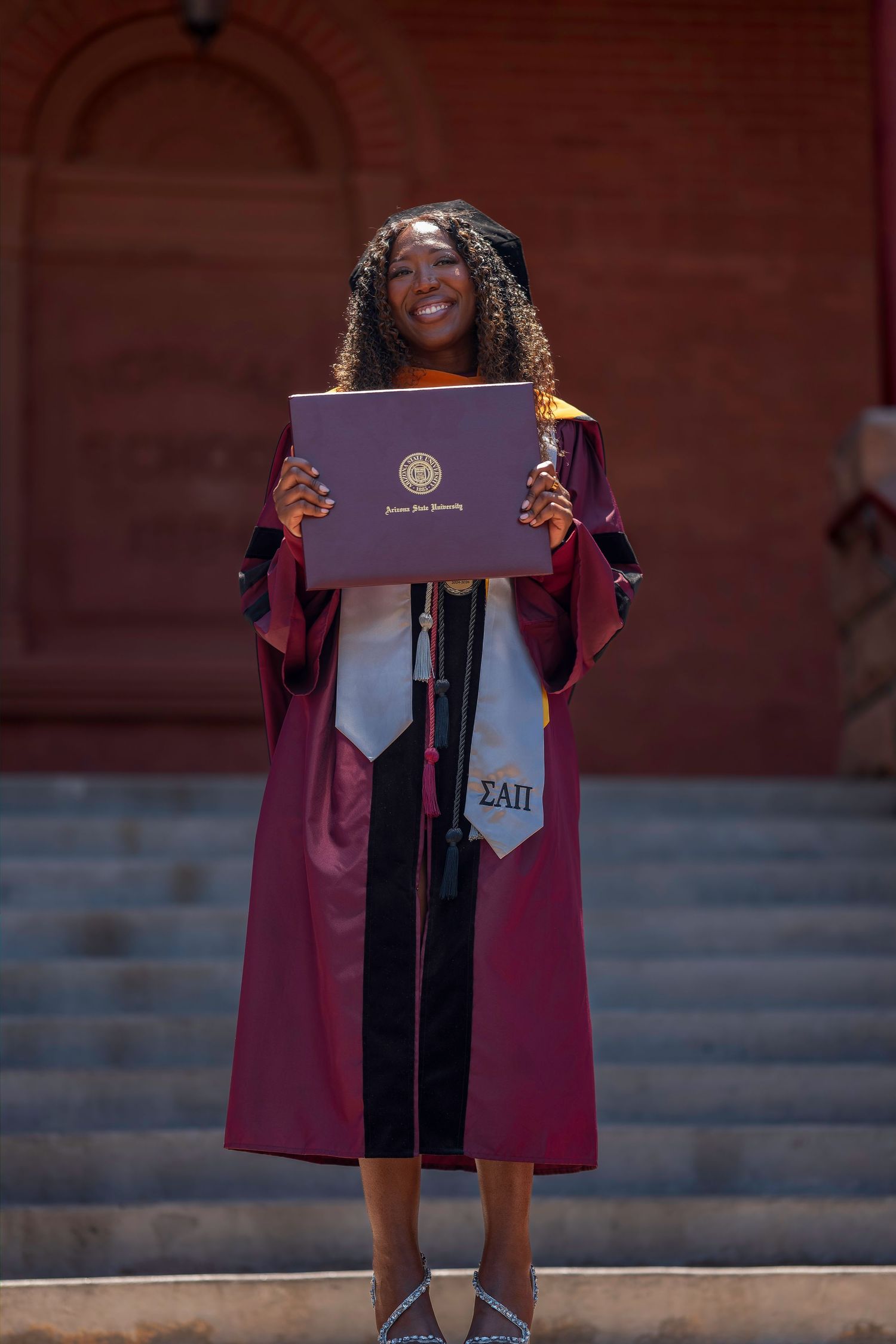 Smiling graduate in maroon cap and gown holds diploma, wearing Sigma Alpha Pi sash on university steps.