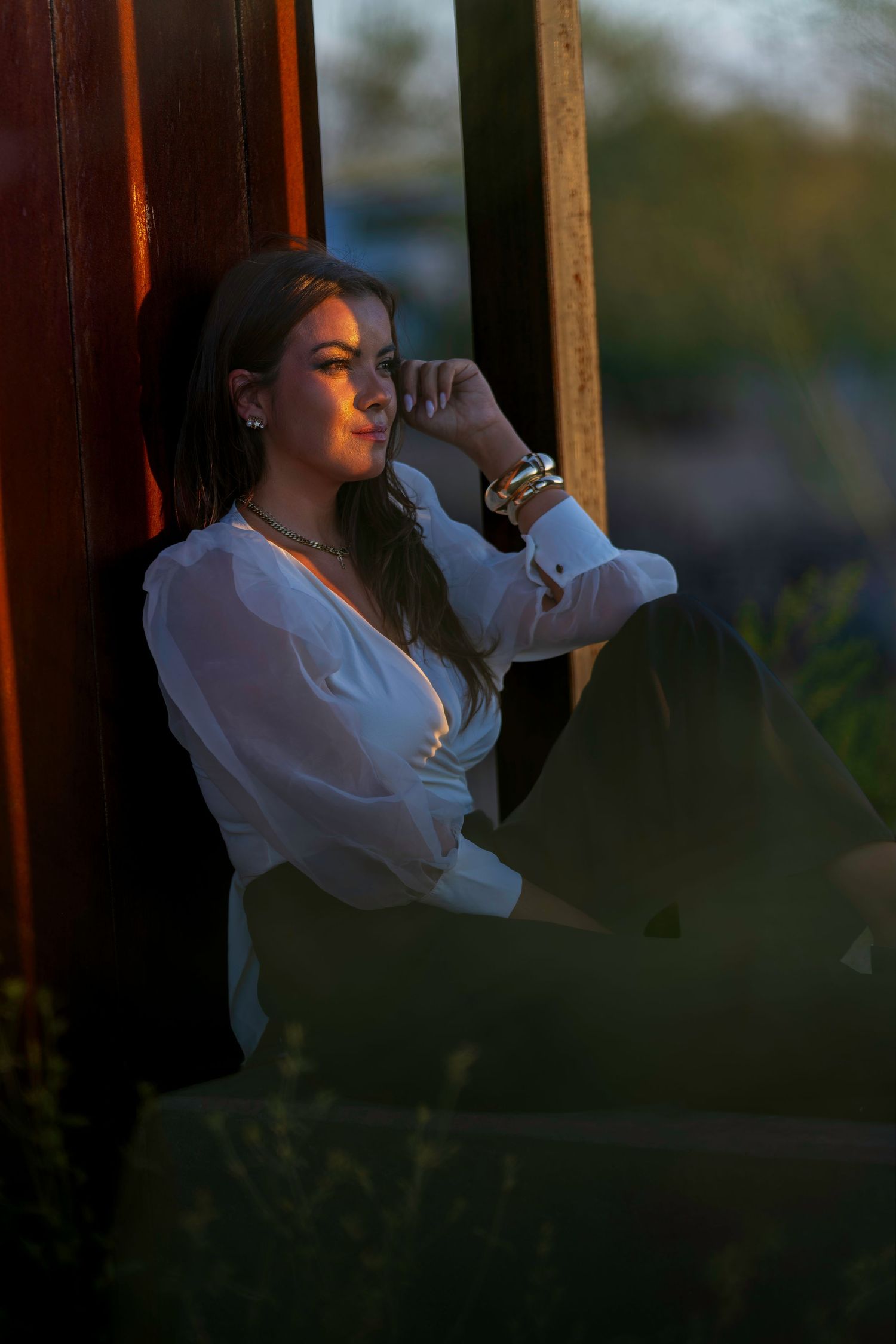 Woman in white blouse leaning against wall in warm golden sunset light outdoors.