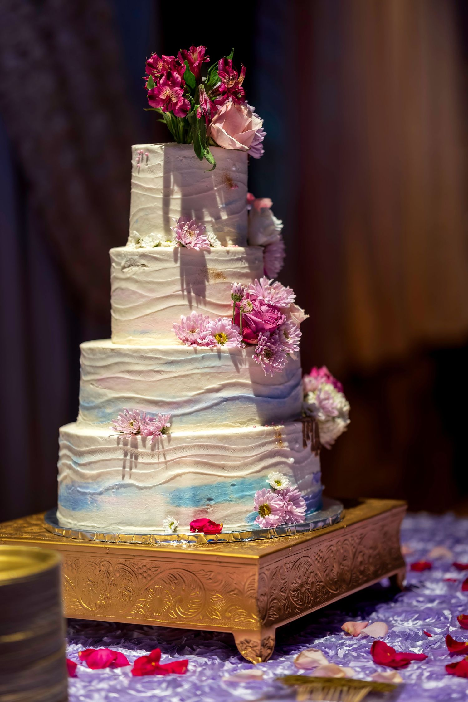 Elegant four-tier white wedding cake with blue watercolor details and pink flowers on a gold stand.