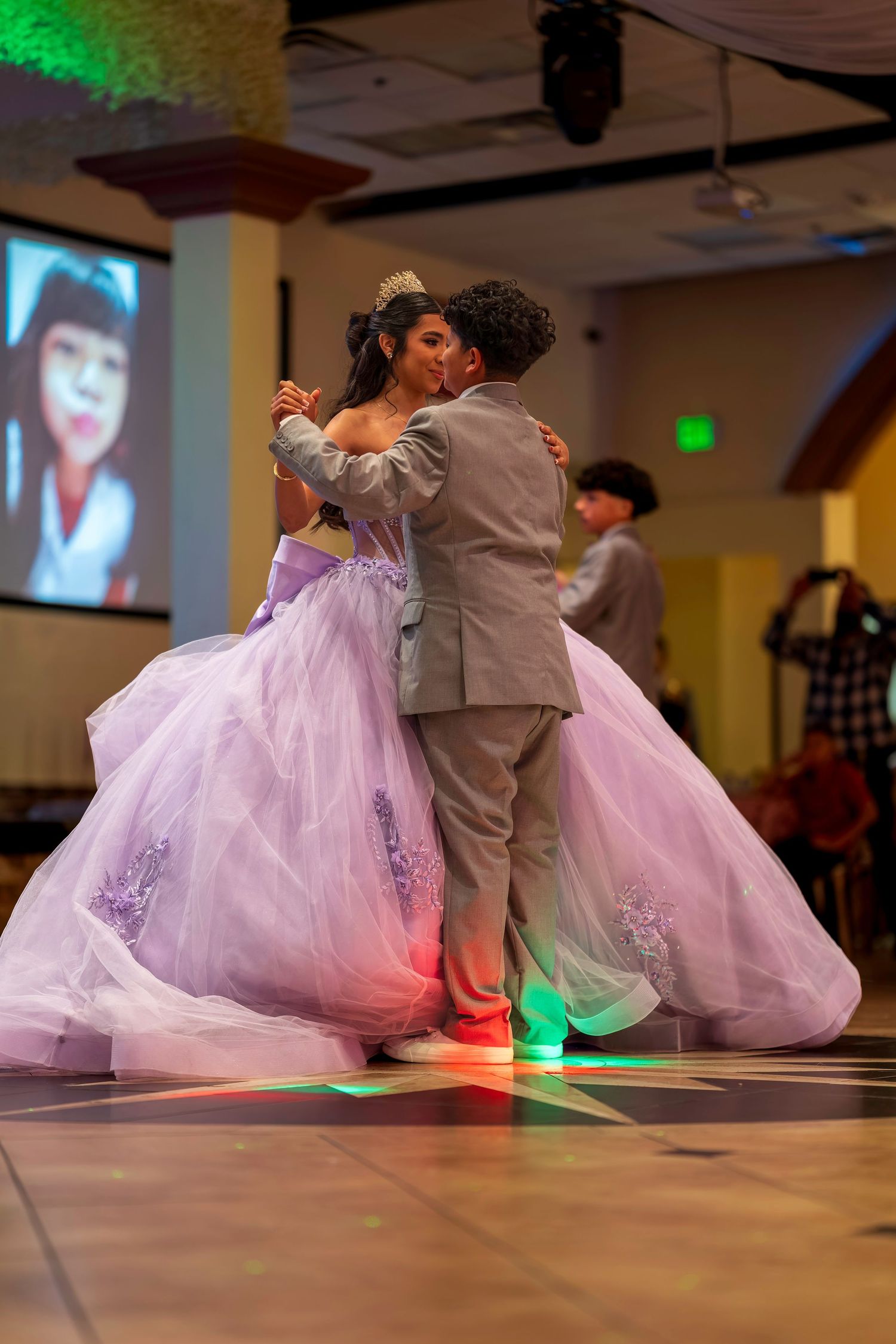 Couple sharing a dance at a quinceañera, wearing a lavender ball gown and gray suit on a colorful dance floor.
