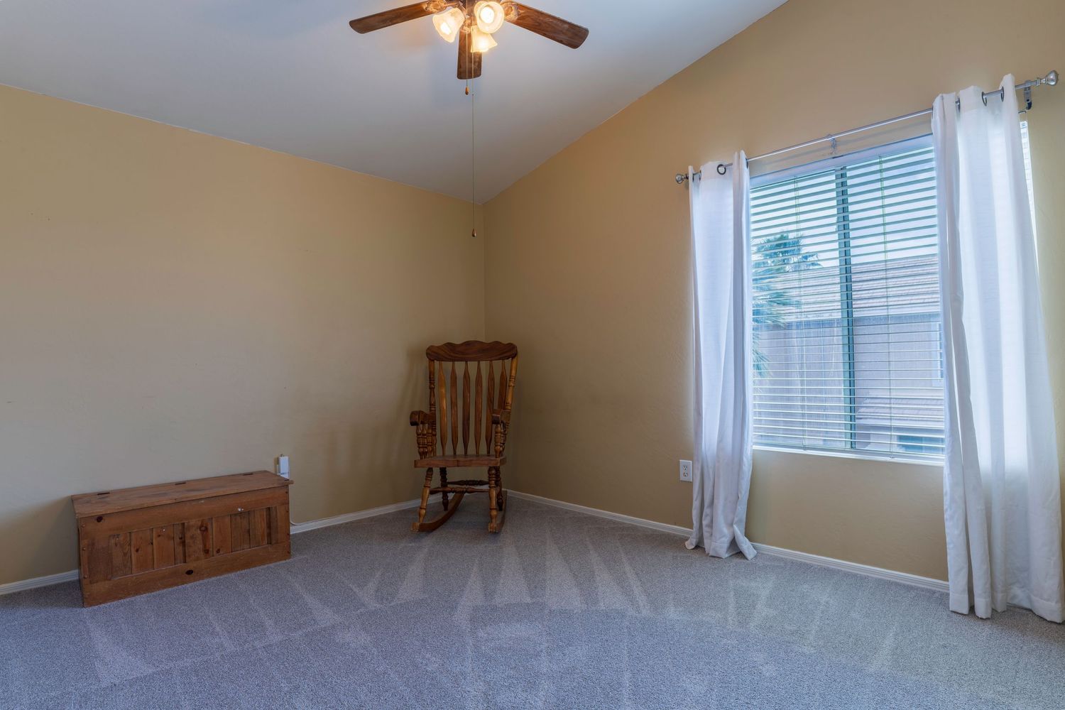 Bright empty bedroom with carpet, ceiling fan, rocking chair, and large window with white curtains.