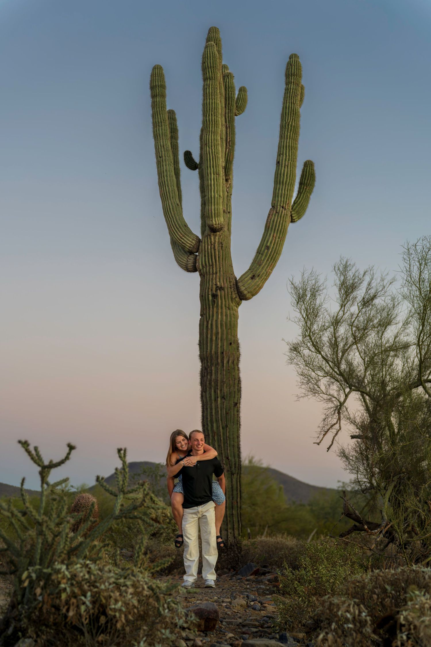 Couple embracing in front of a tall saguaro cactus in the Arizona desert at dusk.
