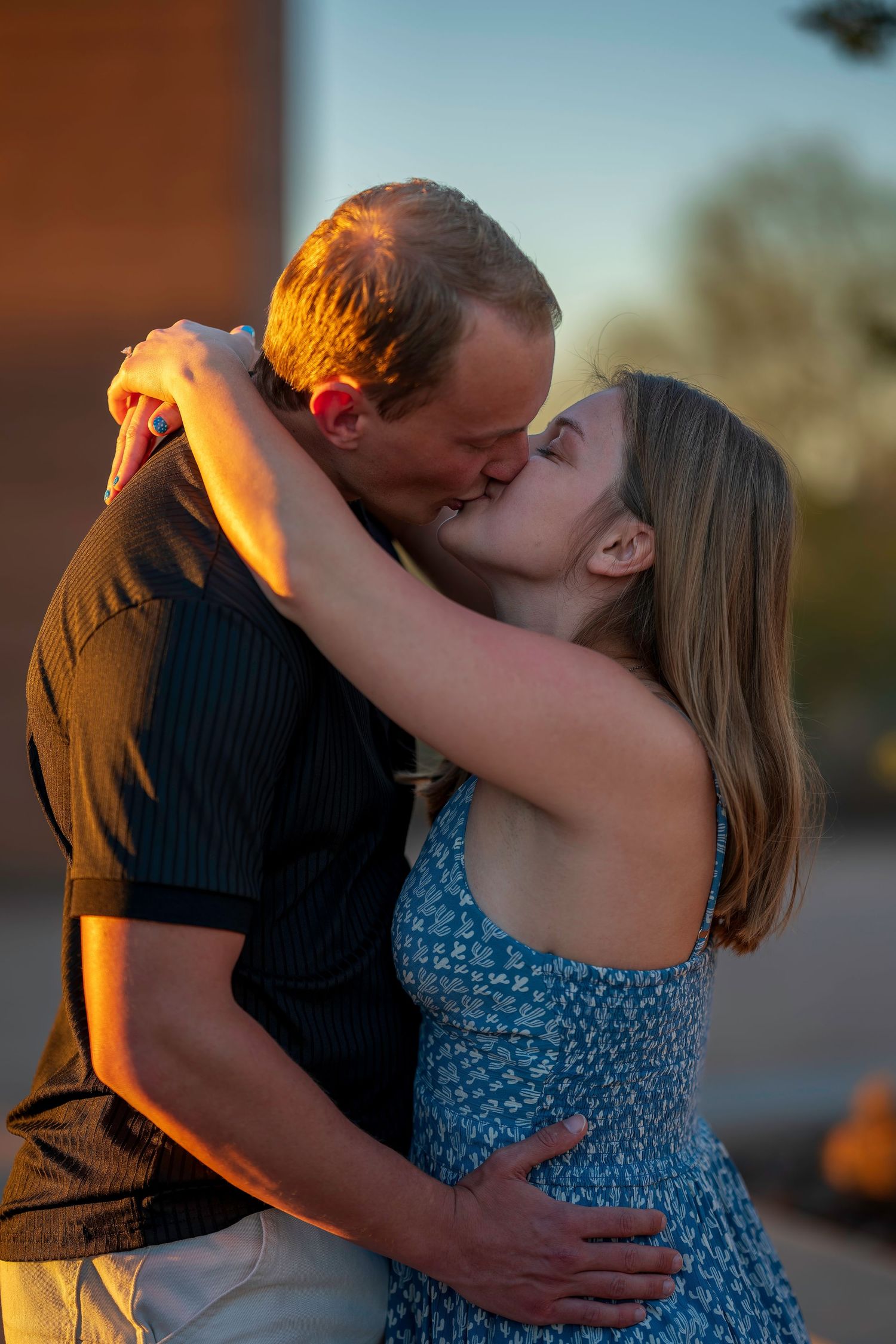 Couple sharing a romantic kiss at sunset, woman in blue floral dress embracing man in black shirt outdoors.