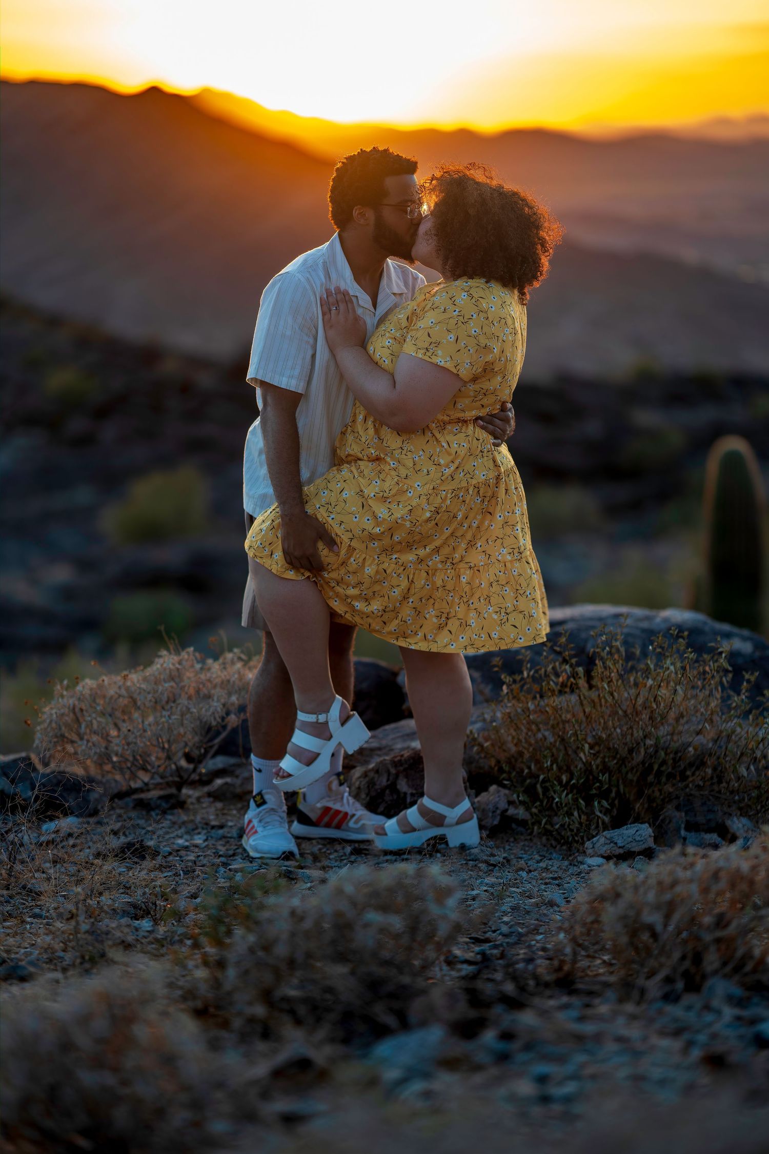 Romantic couple kissing against a vibrant desert sunset with mountains, cacti, and warm golden light.