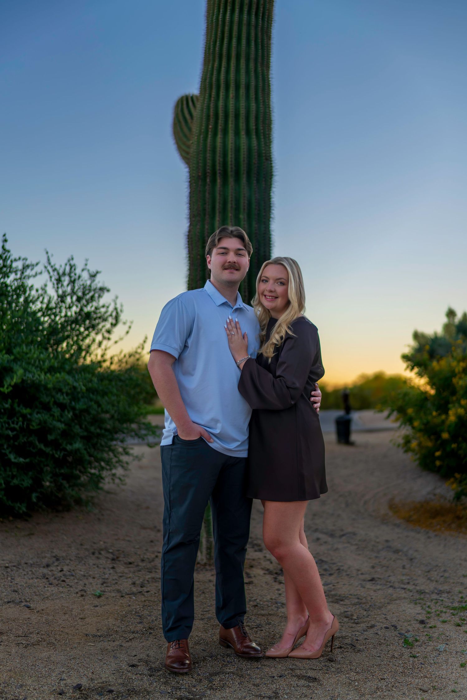 Engaged couple posing together in front of a tall saguaro cactus at sunset in Arizona desert.