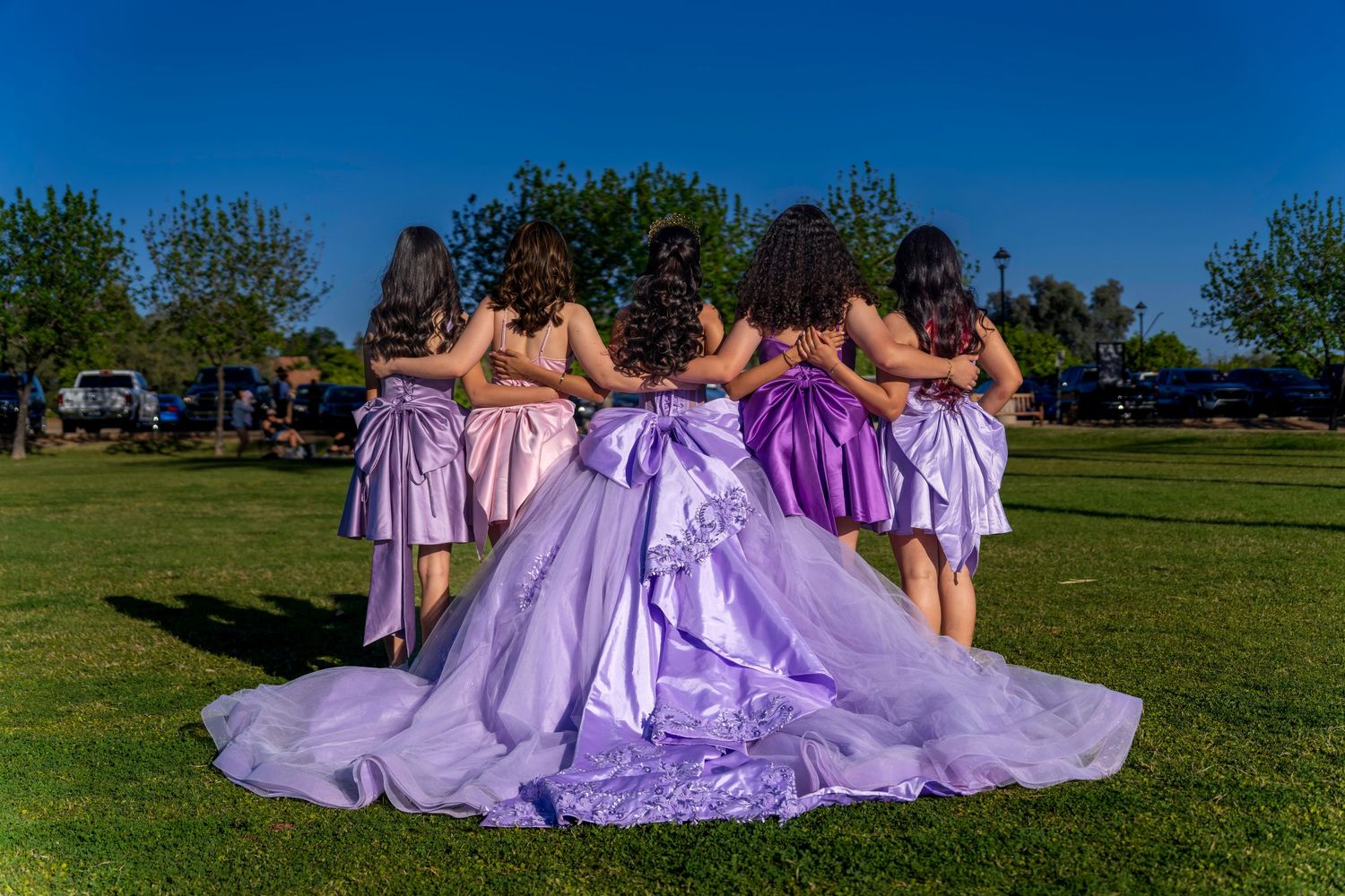 A quinceañera in a lavender ball gown poses with her chambelanes on a green lawn under a clear blue sky.
