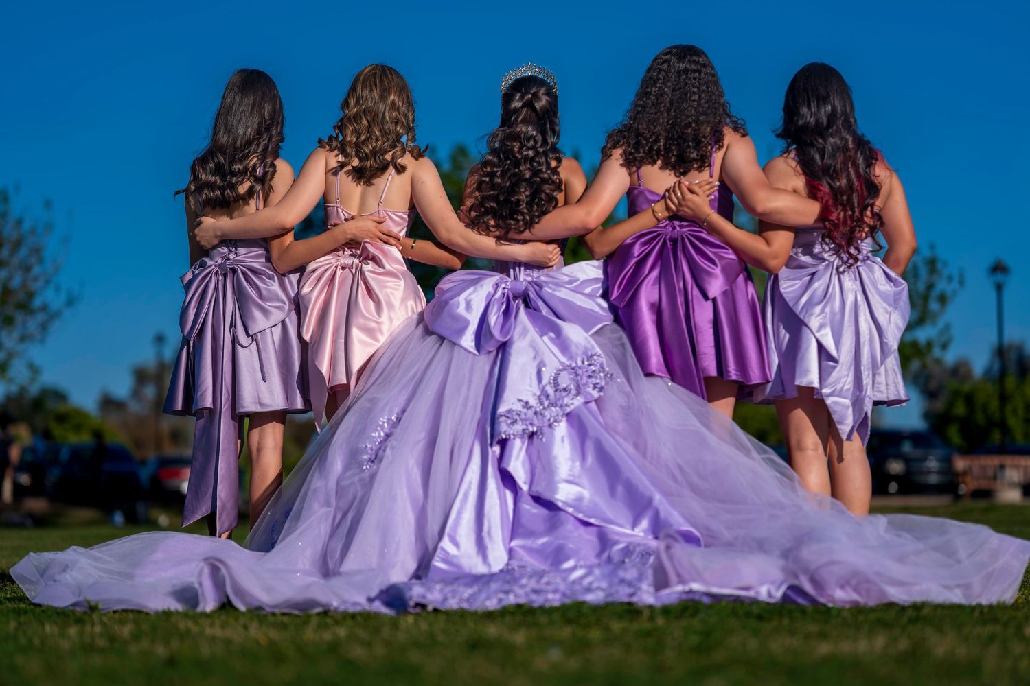 Five young women in elegant satin gowns pose with arms linked, the center girl wearing a lavender quinceañera ballgown.