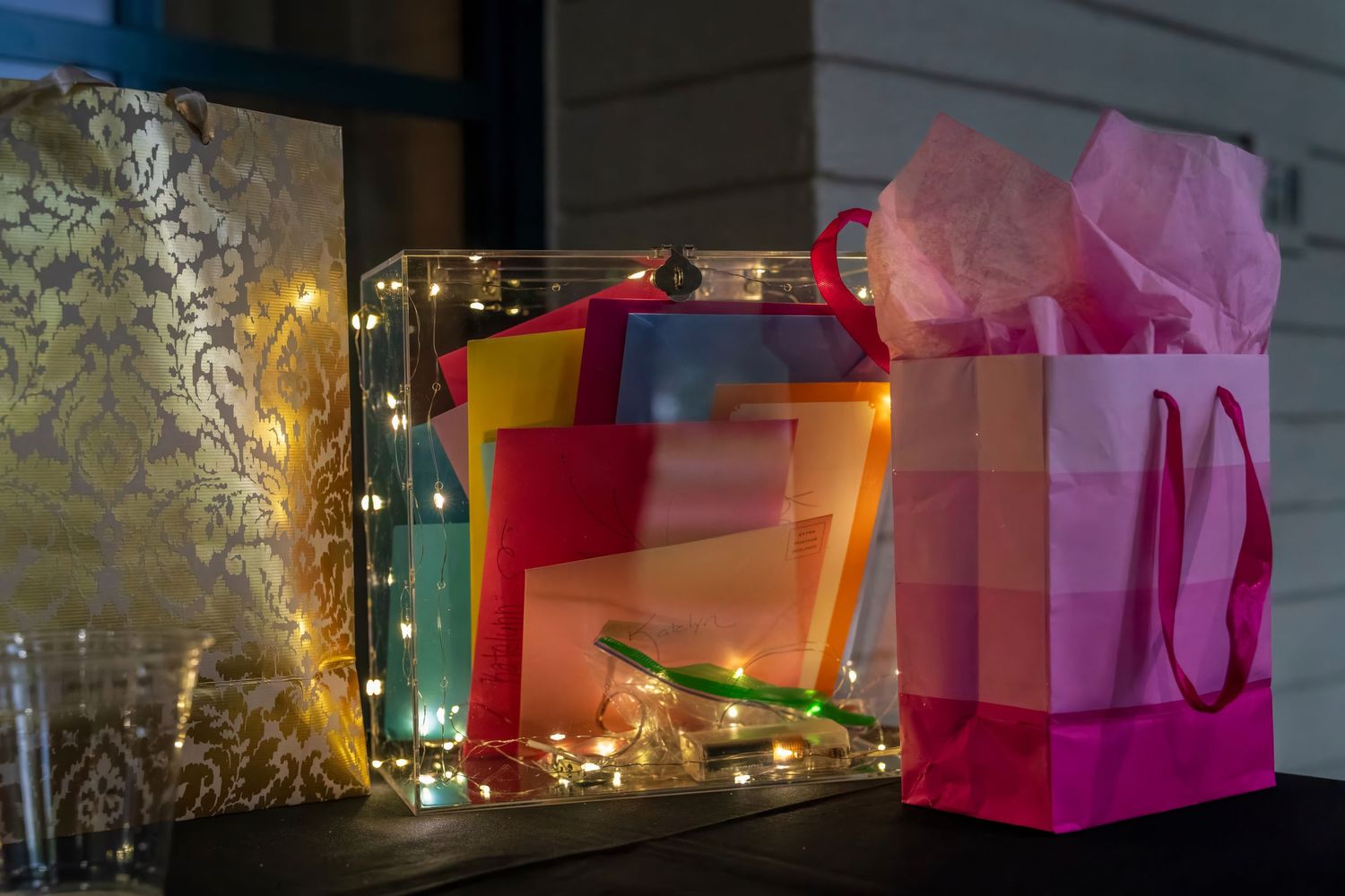 Colorful gift bags and boxes adorned with warm fairy lights arranged on a dark table at a festive event.