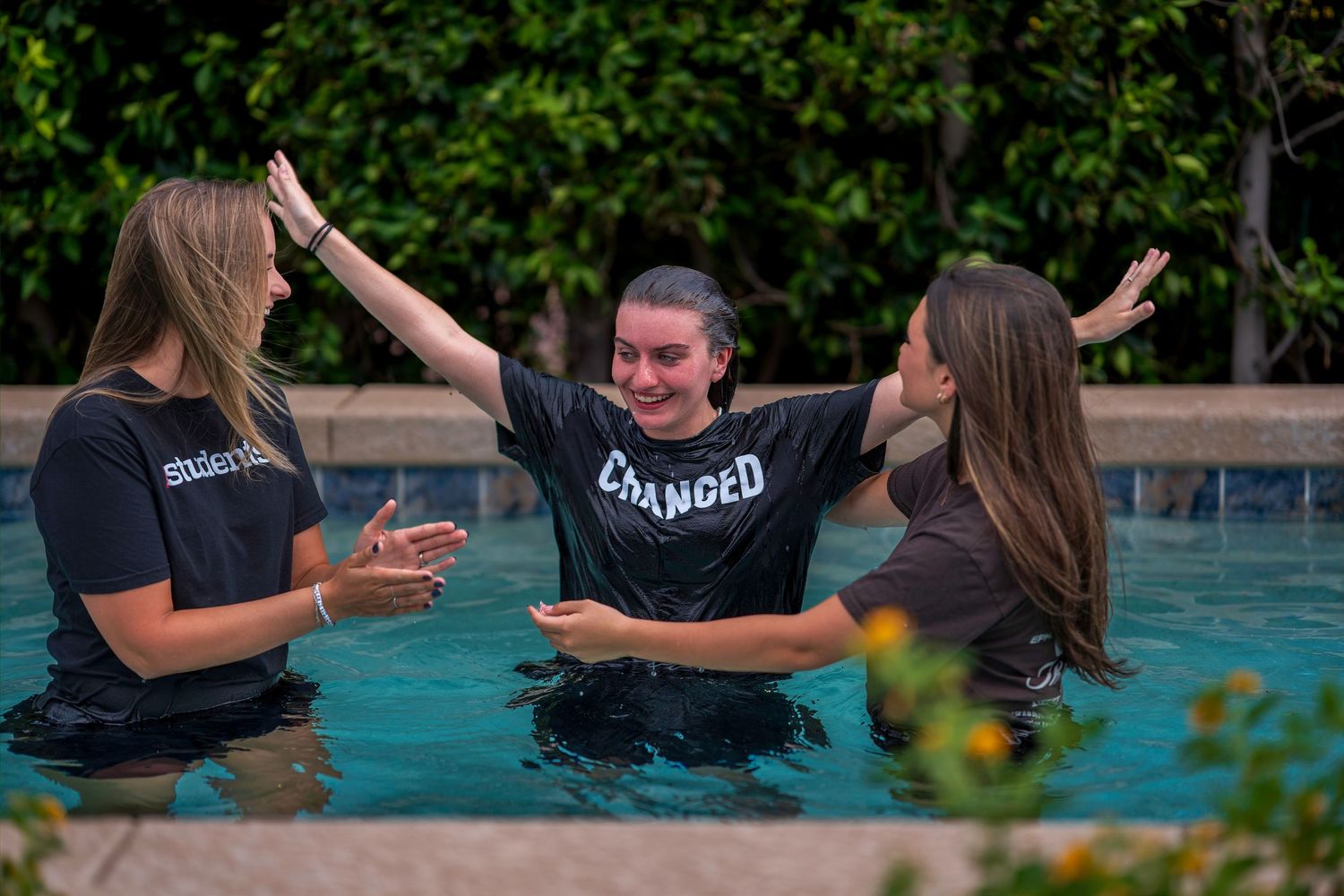 Three young women in black t-shirts celebrating a baptism in a pool, with arms raised in joy.