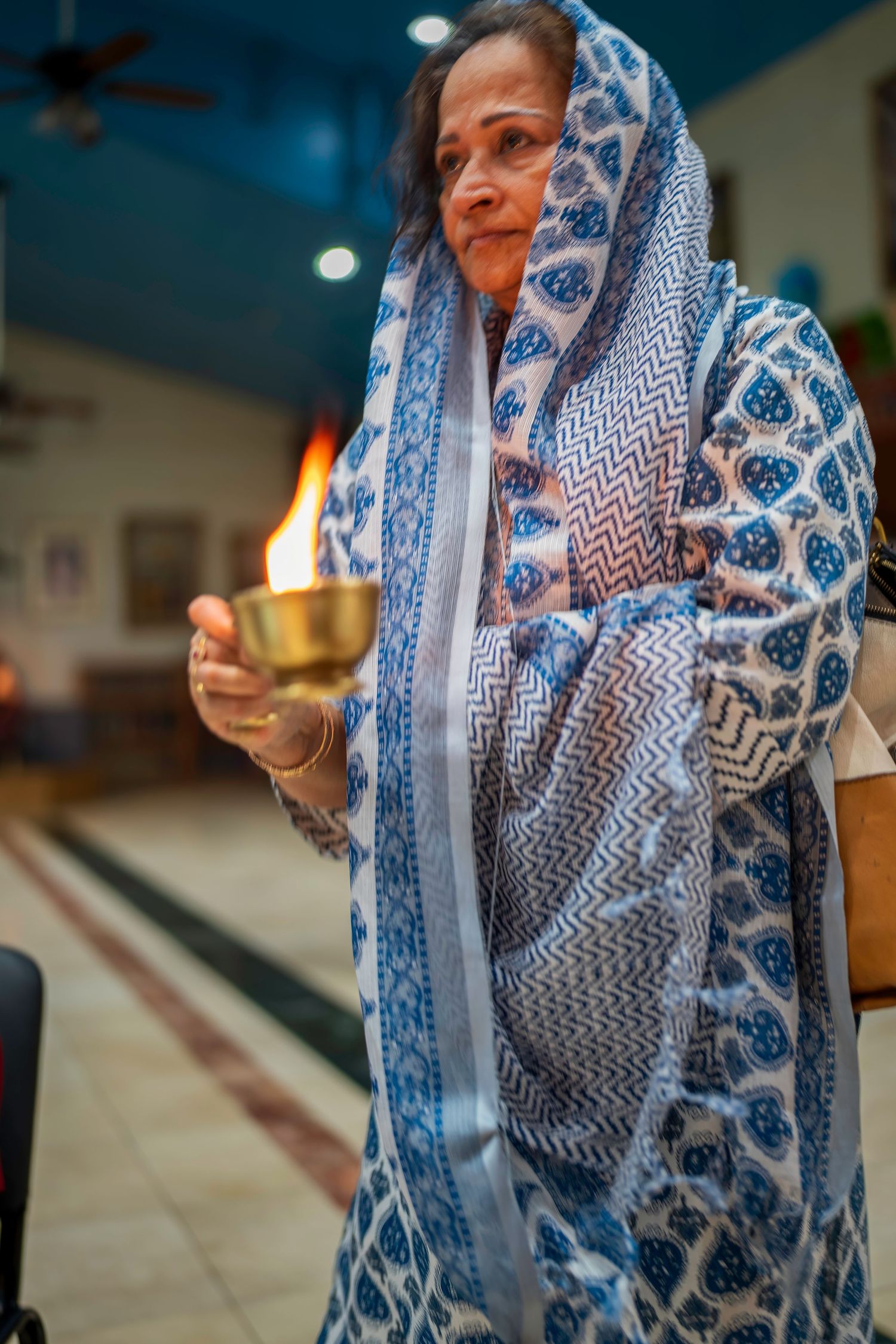 Indian woman in blue floral dupatta holding brass diya lamp during Hindu prayer ceremony indoors.