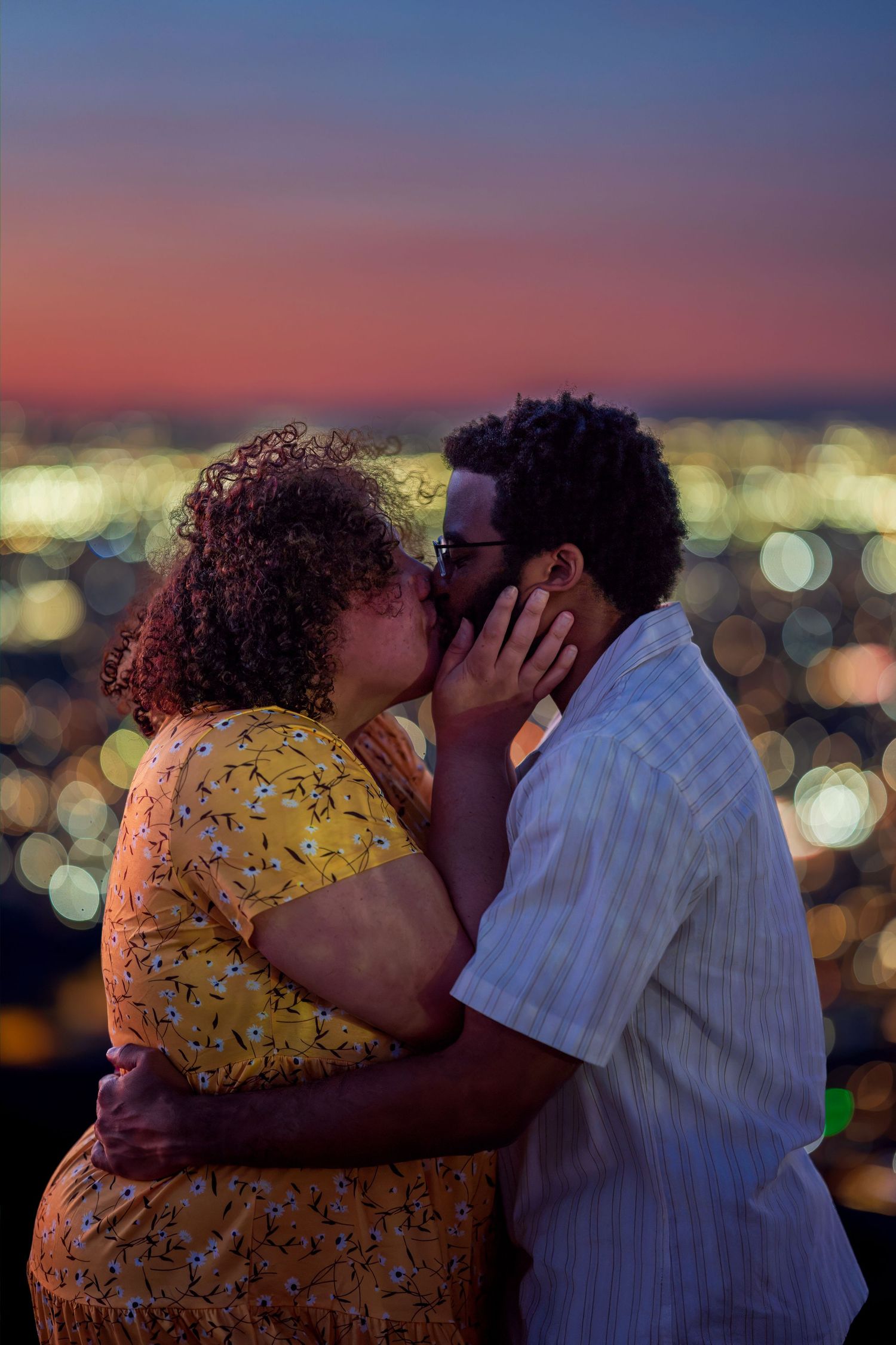 Couple sharing a passionate kiss at night with a glowing city skyline bokeh background behind them.