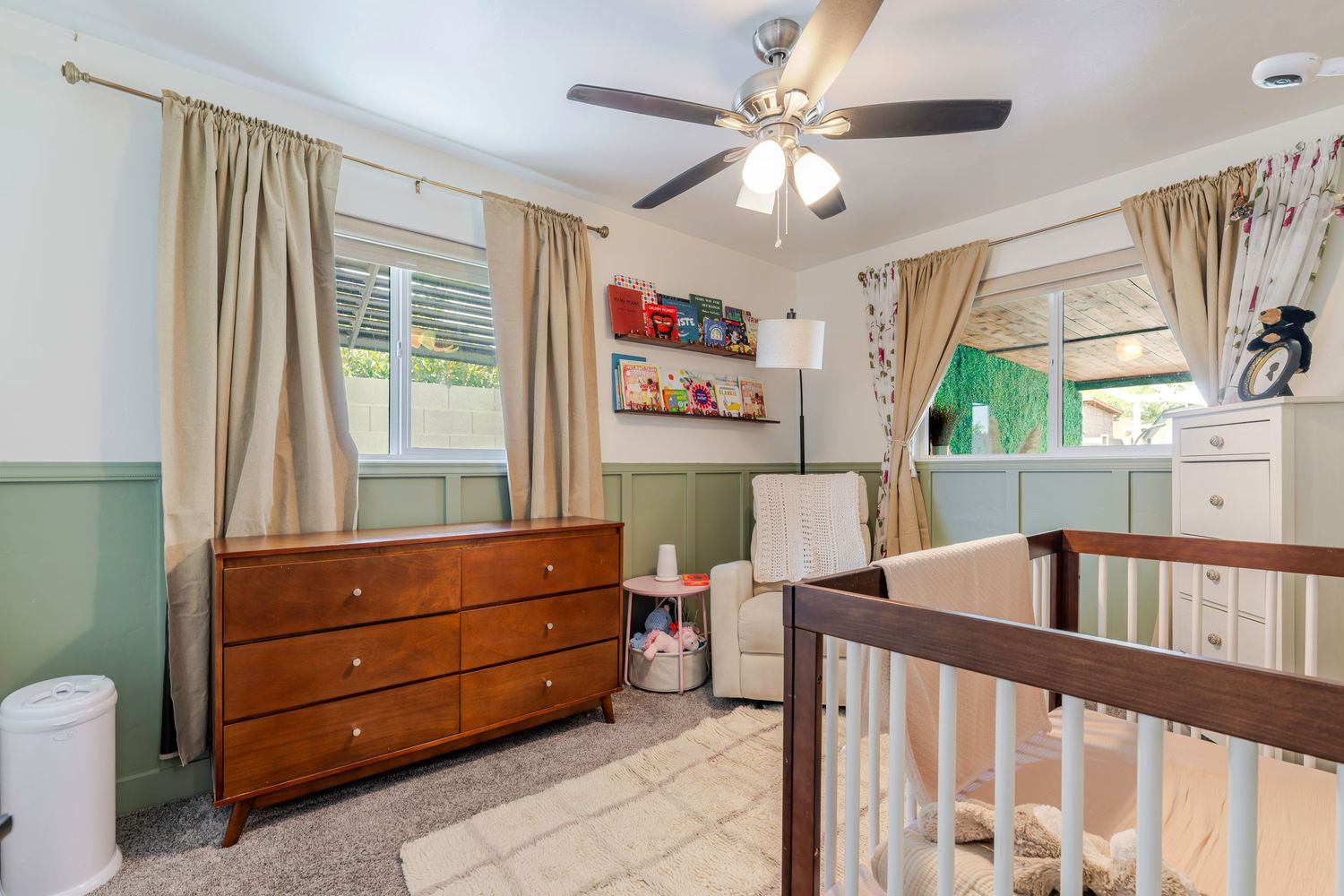 Cozy nursery with wooden crib, dresser, ceiling fan, green accent walls, and colorful curtains in natural light.