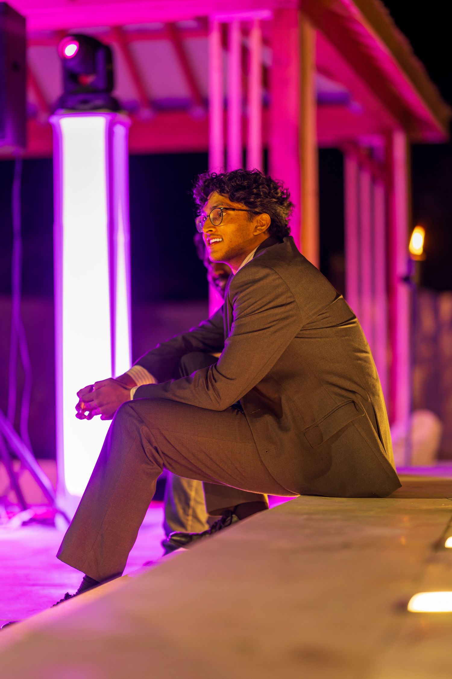 A person in a suit jacket sits on a ledge illuminated by vibrant purple lighting.