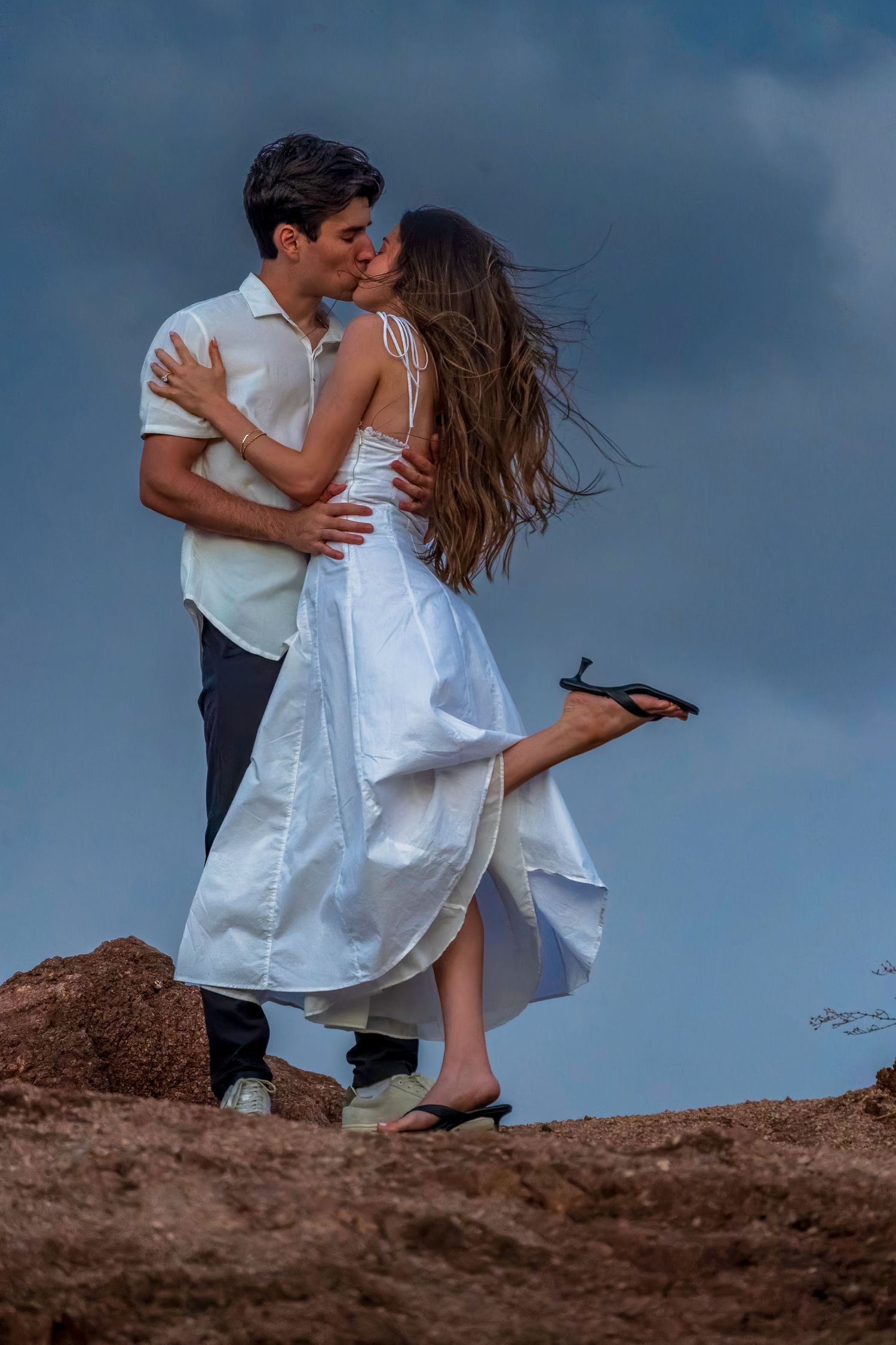 Couple sharing romantic moment on hilltop with dramatic sky background.