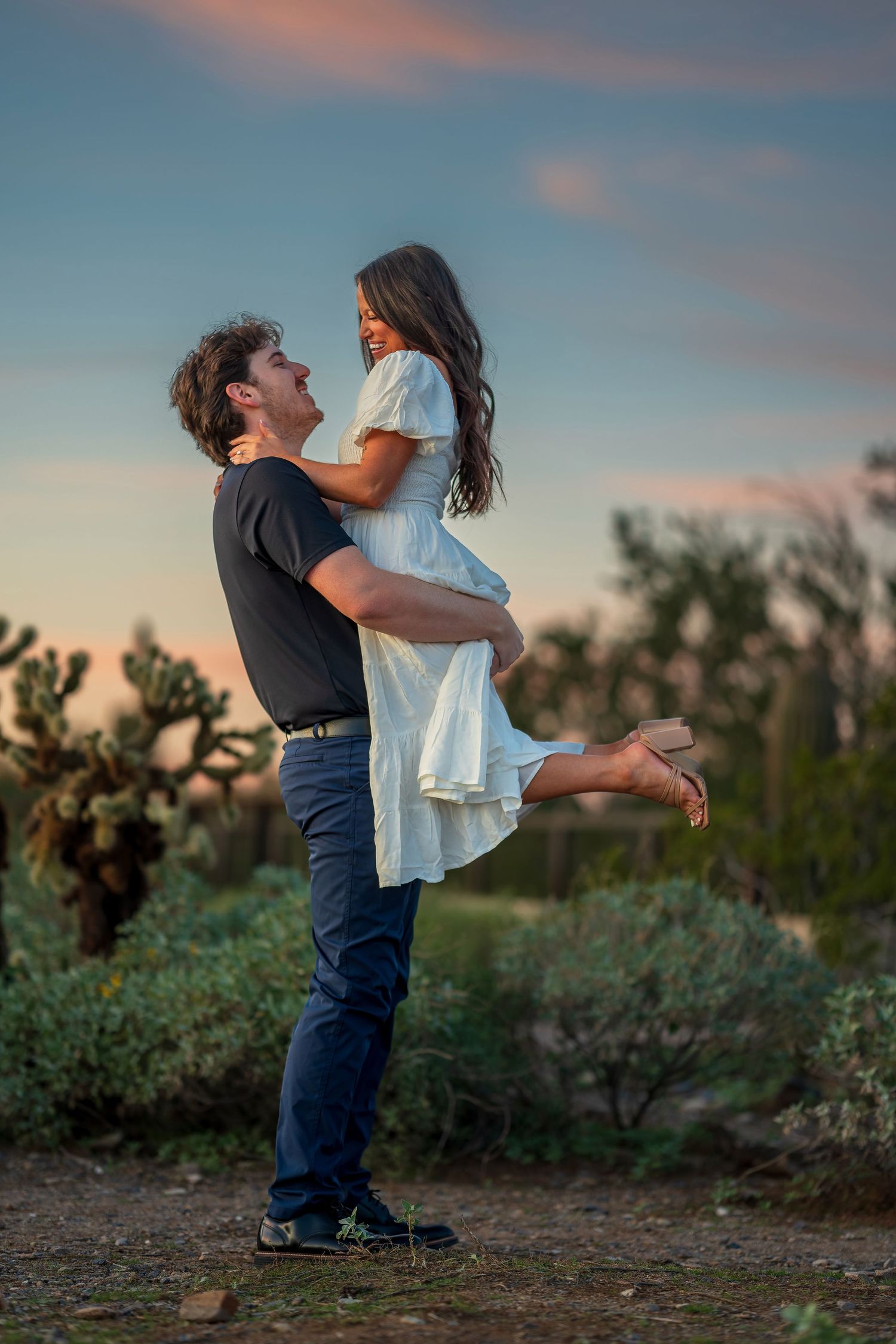 A joyful couple celebrates their engagement with a playful lift in the desert at sunset.