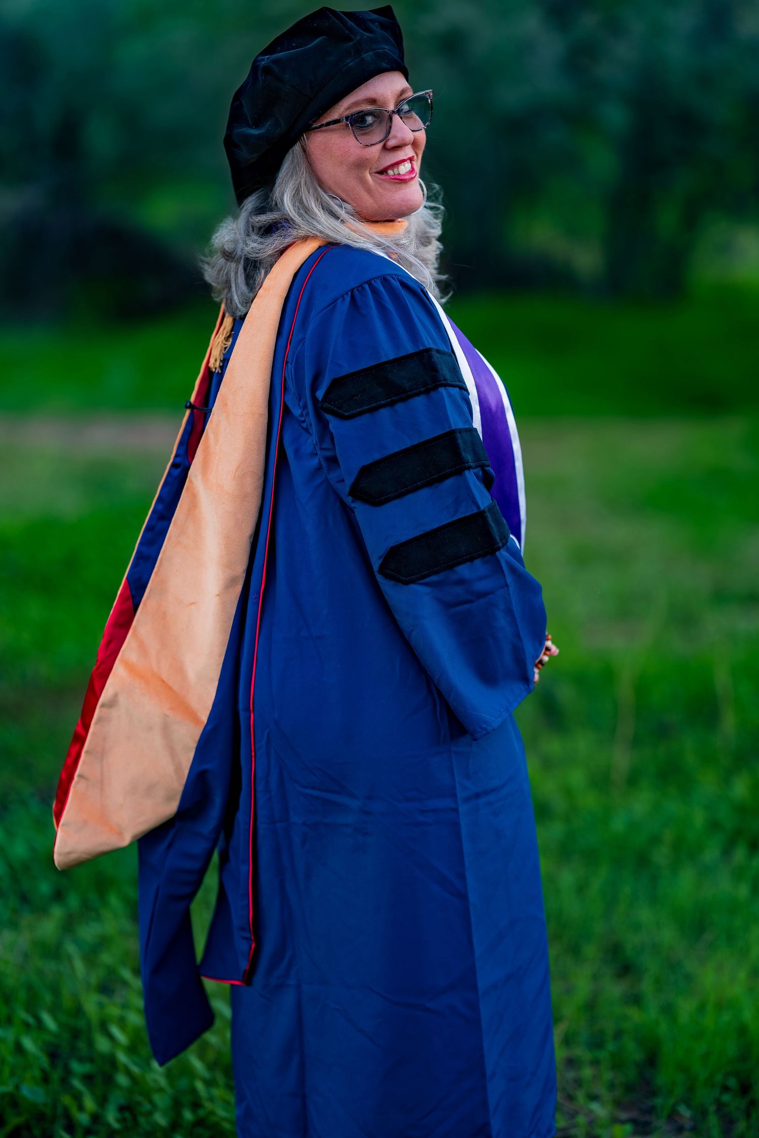 Academic wearing blue doctoral graduation gown poses outdoors with orange and purple stoles.
