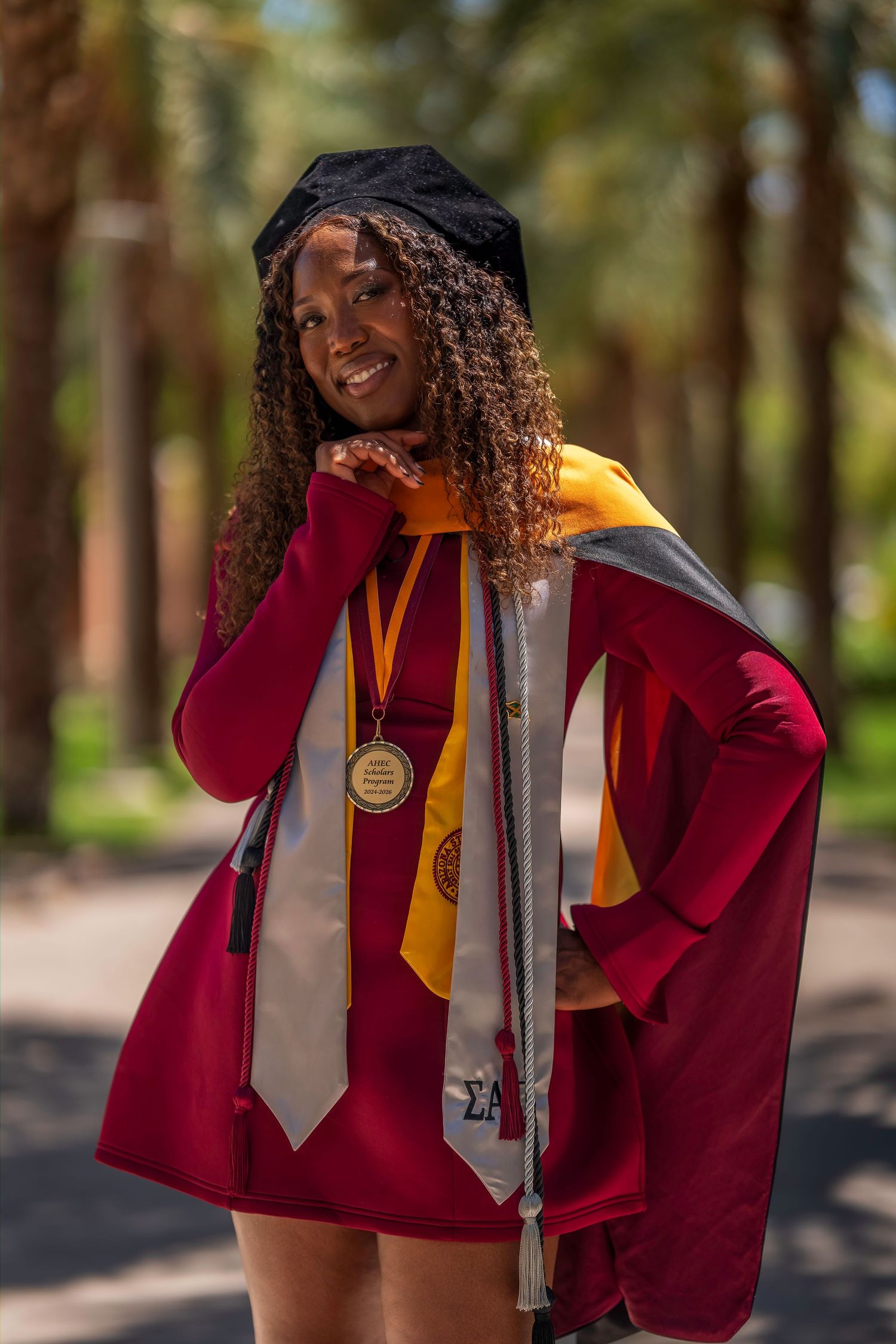 Smiling ASU graduate in maroon cap and gown with Sigma sorority stole and achievement medal.