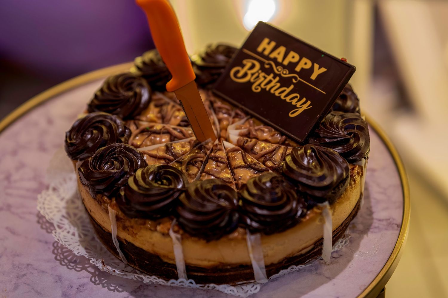 Chocolate birthday cake with decorative rosettes, Happy Birthday sign, and lit candle on a elegant plate.