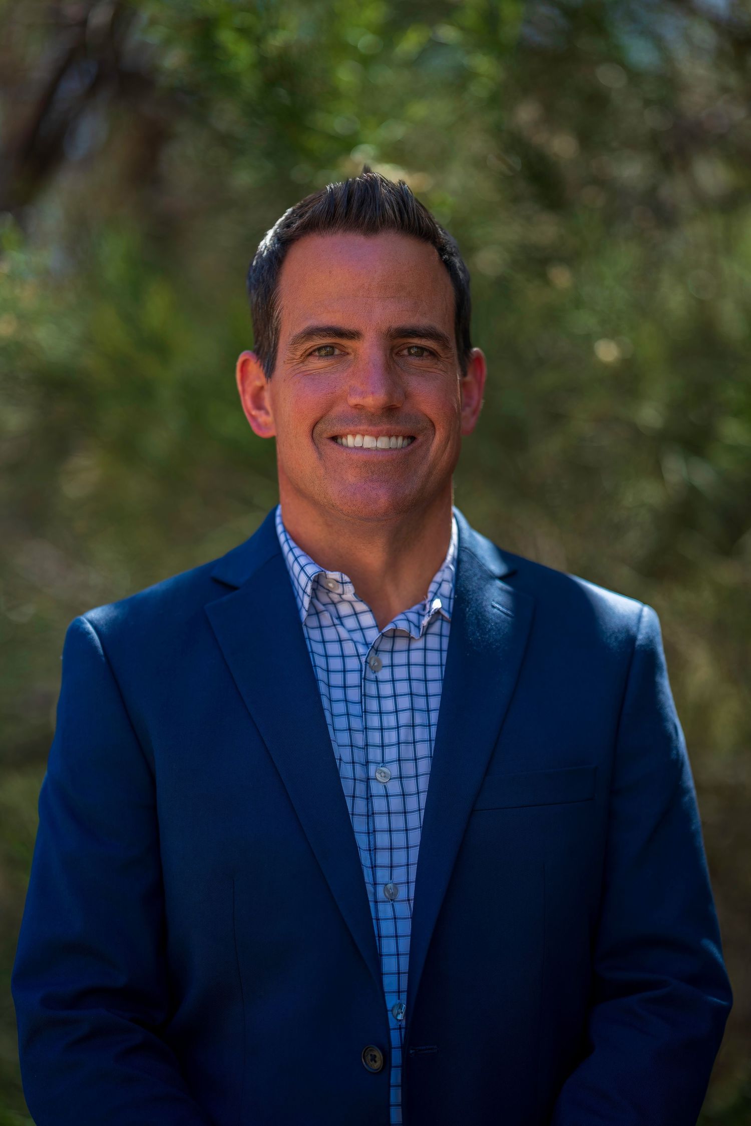 Smiling man in blue blazer and checkered shirt poses outdoors with green trees in background.