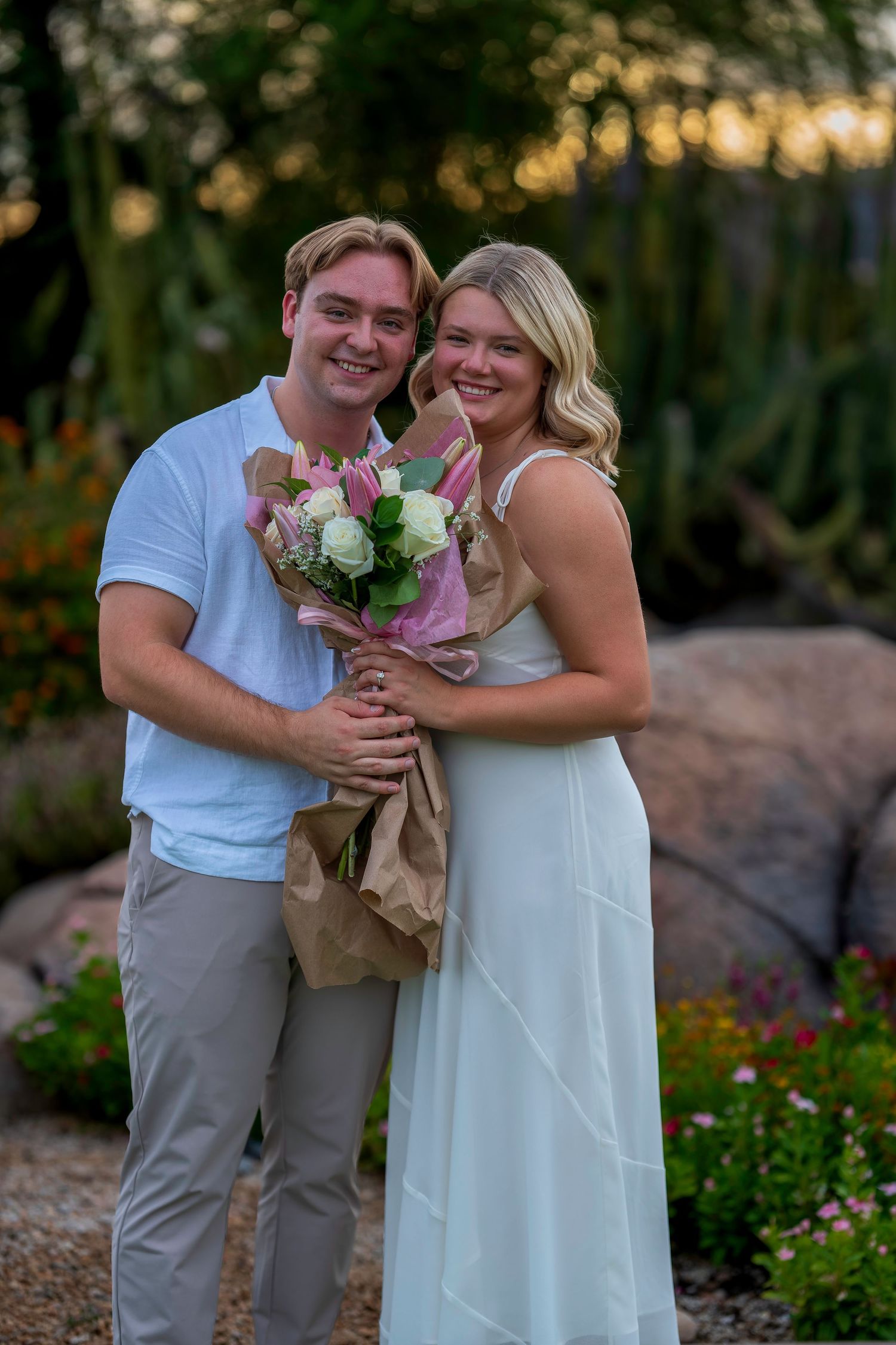 Smiling couple holding pink and white flower bouquet in garden at sunset, man in white shirt, woman in white dress.