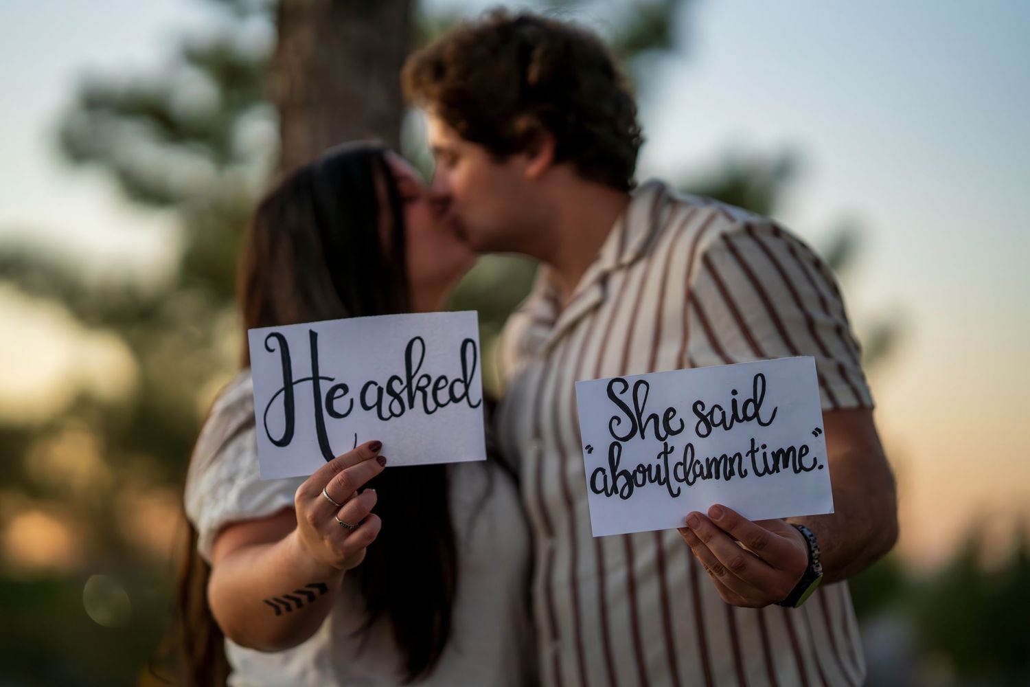 Newly engaged couple kissing while holding signs reading He asked and She said about damn time.