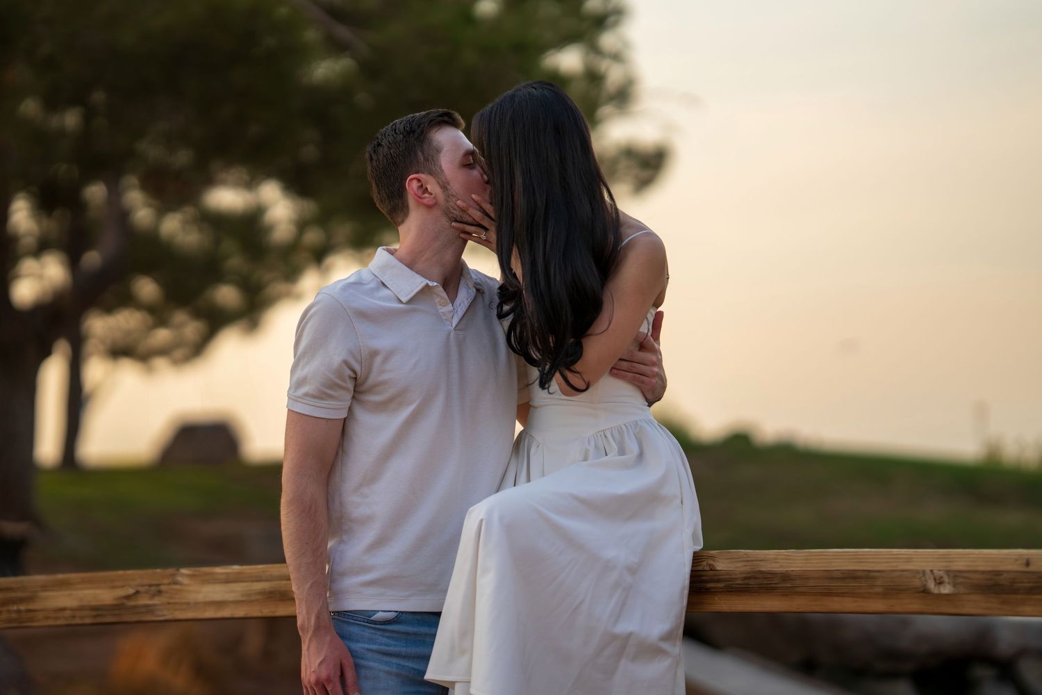 Couple sharing a romantic kiss at sunset on a wooden fence in an outdoor setting.