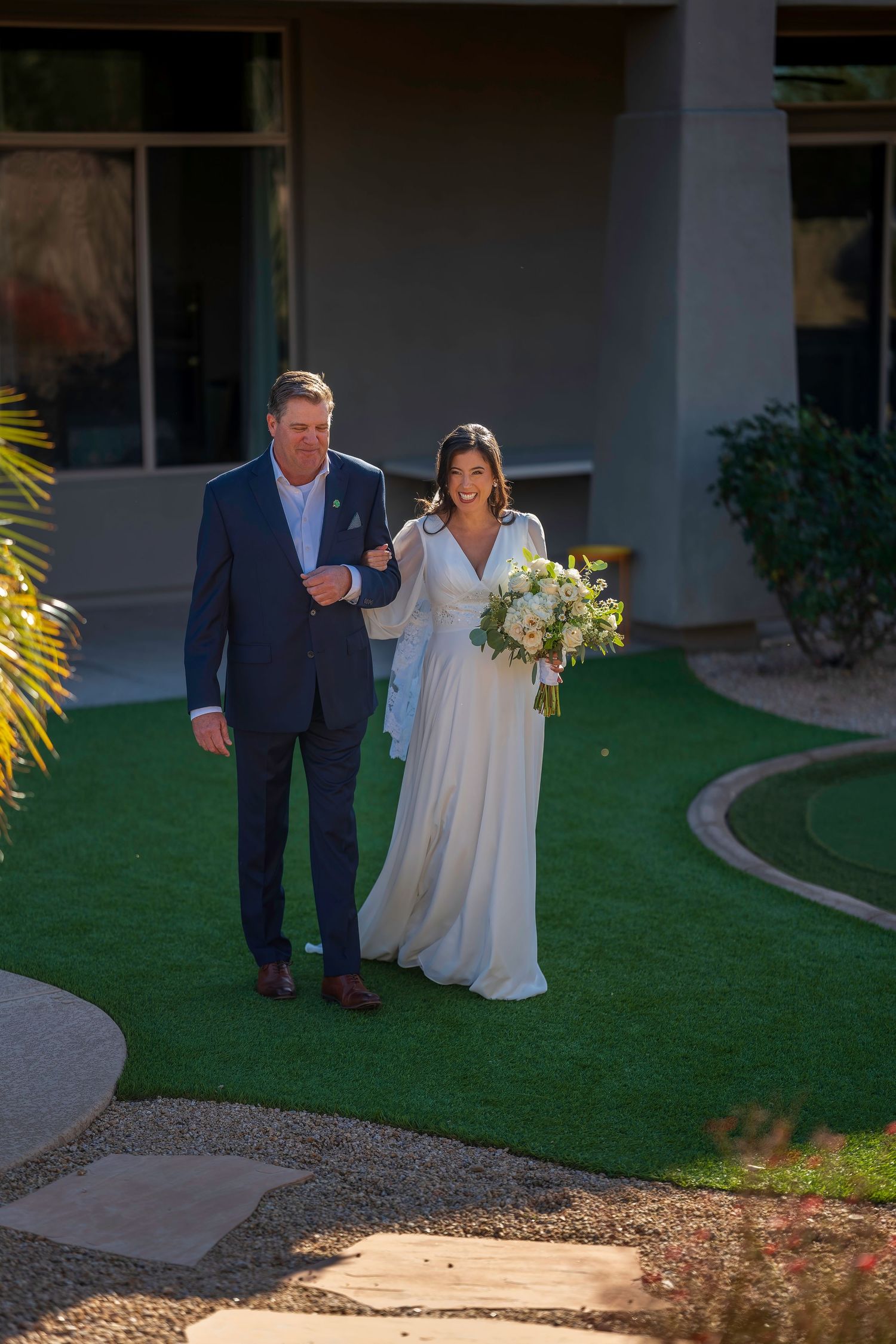 Bride in white flowing gown holding bouquet walks arm-in-arm with man in navy suit on green lawn.