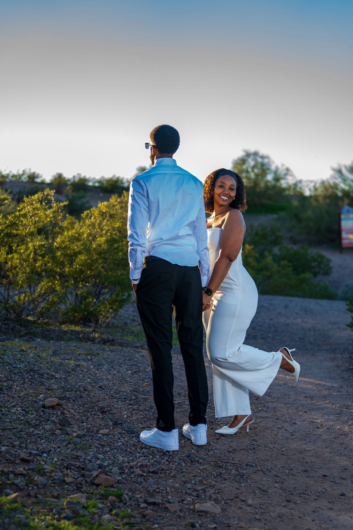 A smiling bride in white dress holds her grooms hand while walking on a scenic desert trail.