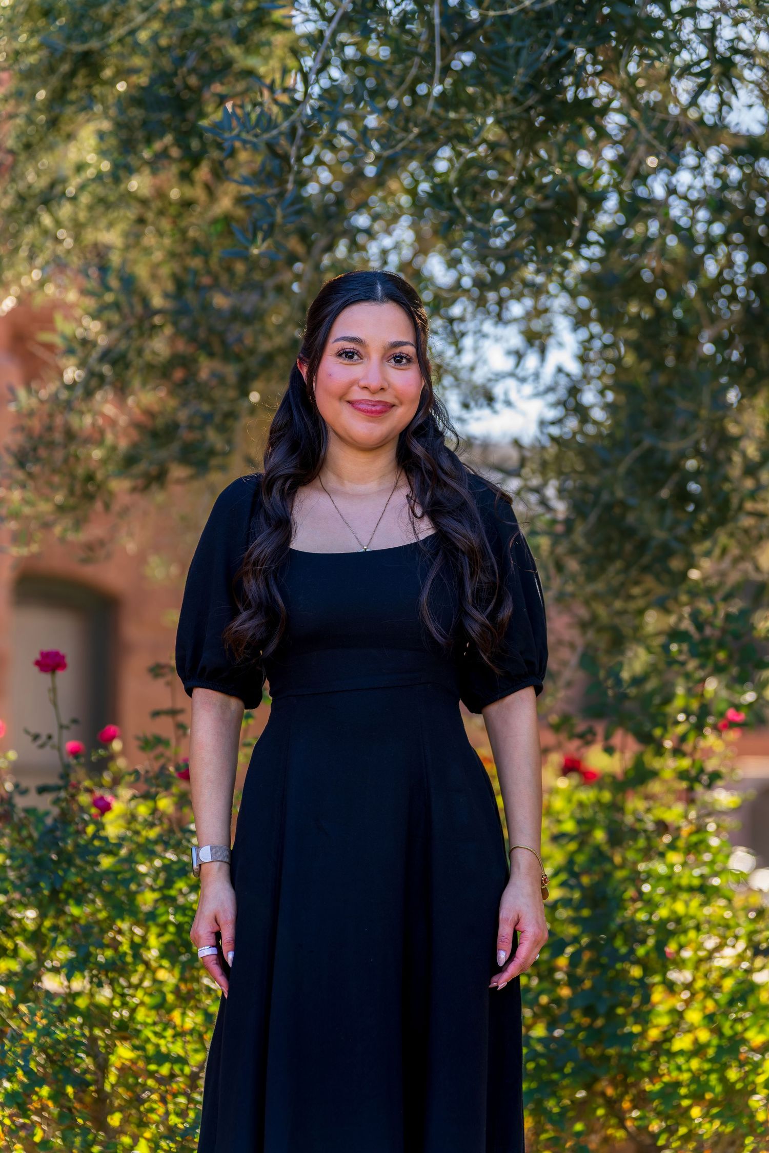 Portrait in black dress against garden backdrop with flowering bushes and brick architecture.