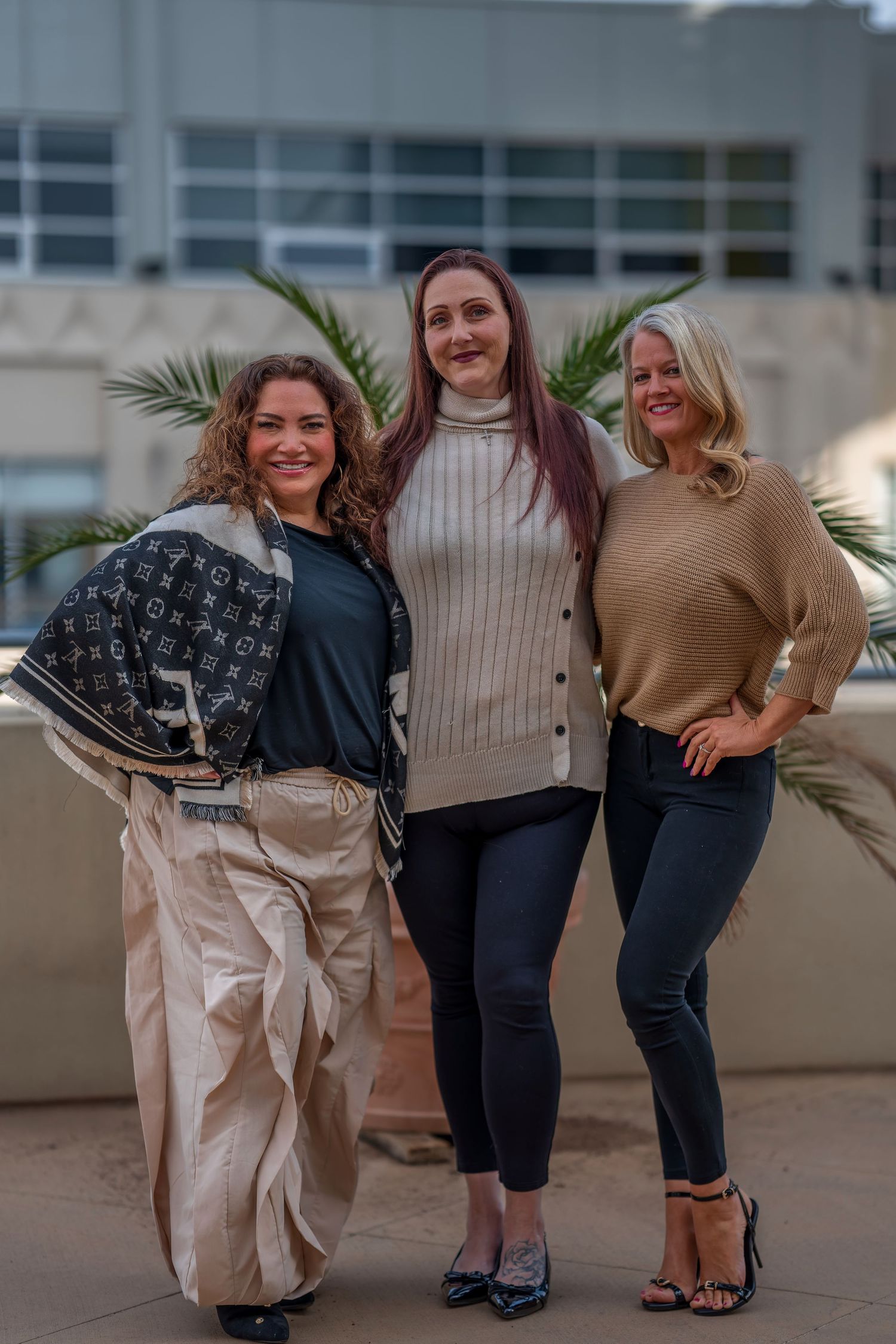 Group posing casually outdoors wearing neutral toned outfits near potted palm plants.