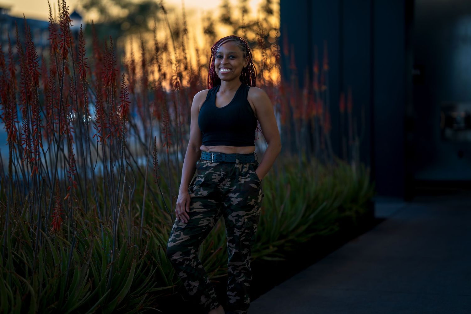 Outdoor photo shoot at dusk with camouflage pants and black top against natural foliage.