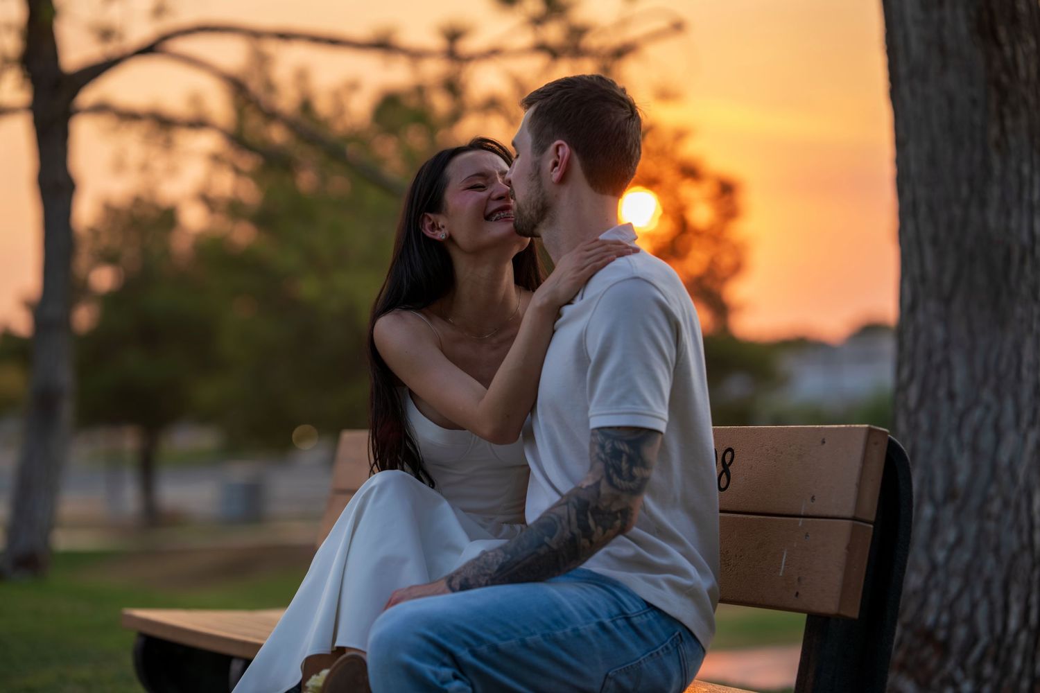 Engaged couple sitting on park bench sharing a kiss during golden sunset, man has tattoos.
