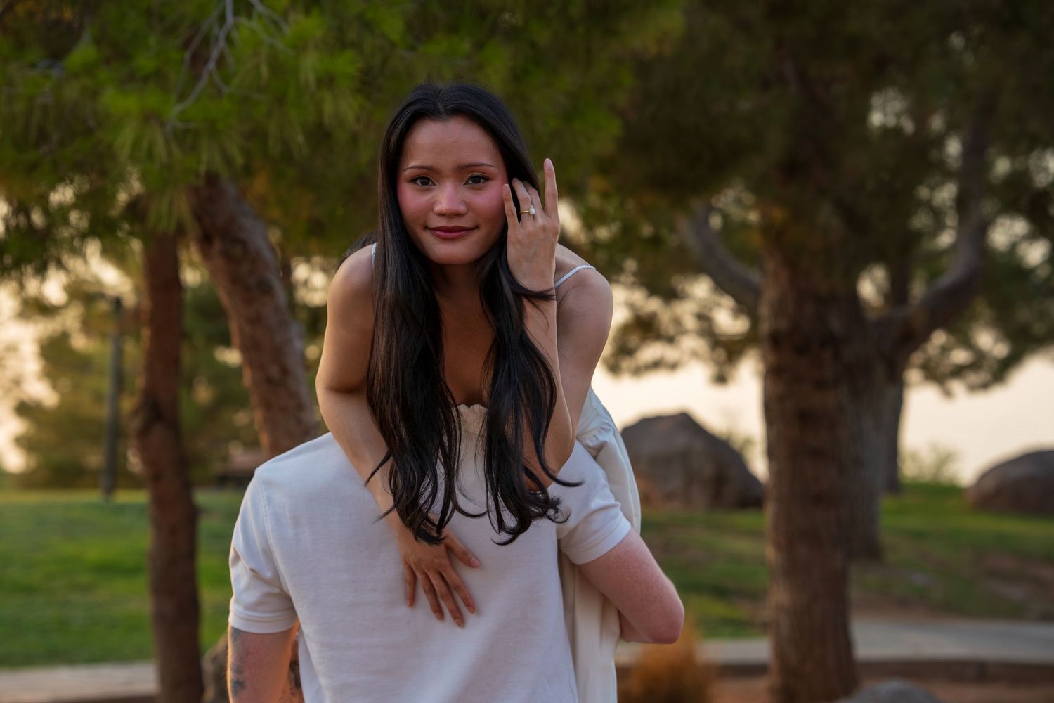 Young woman with long dark hair smiling while getting a piggyback ride in a sunny park.