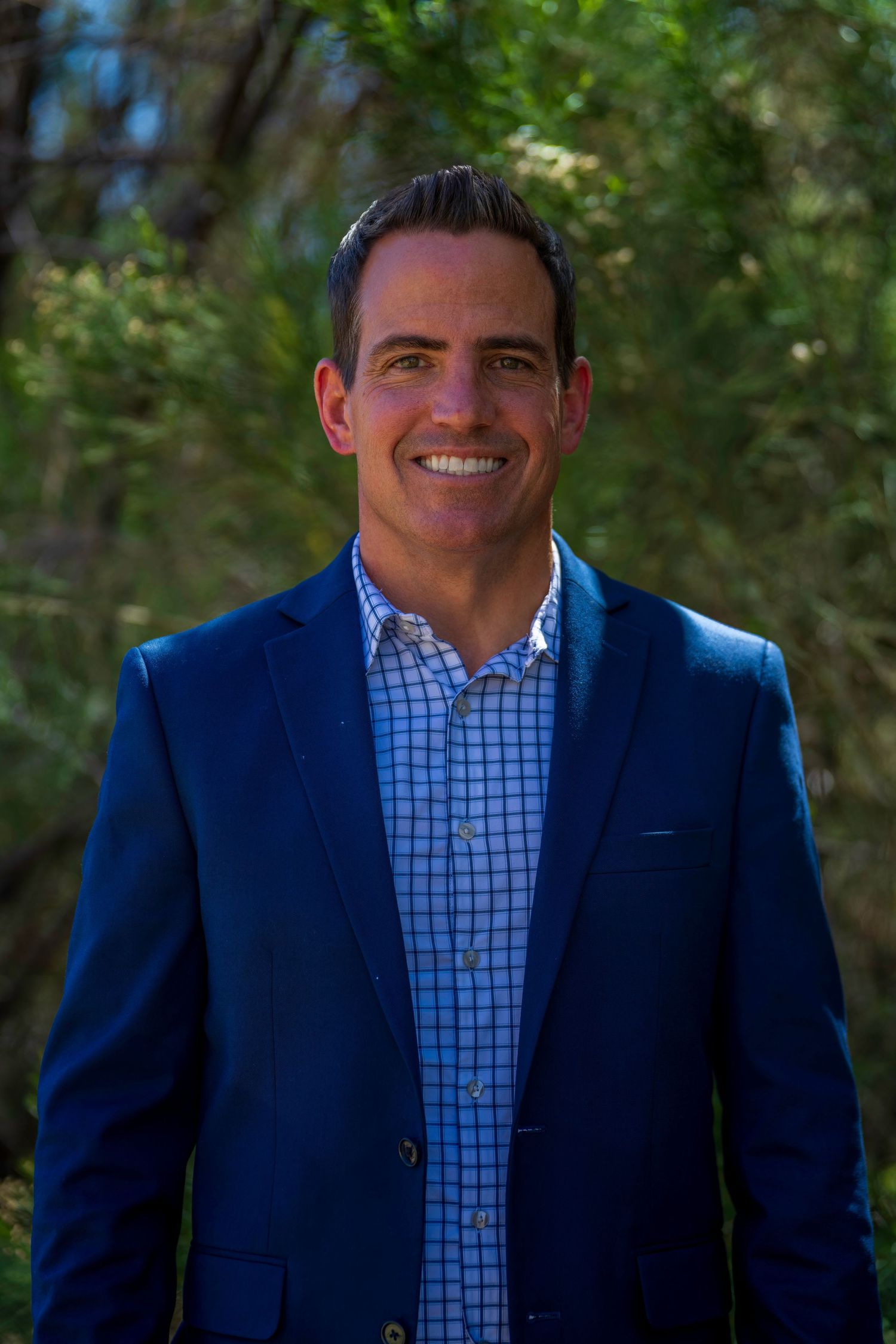 Smiling man in blue blazer and checkered shirt poses outdoors with lush green trees in background.