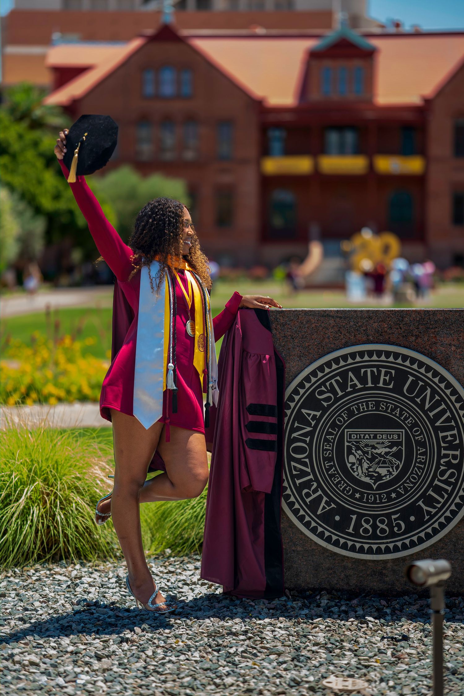 ASU graduate in maroon cap and gown posing by Arizona State University 1885 stone campus sign.