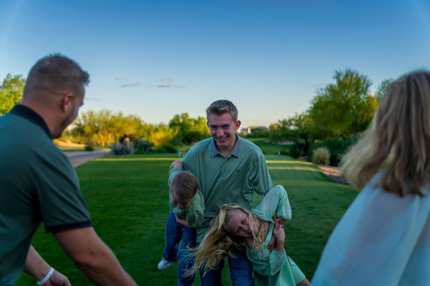 Family playing outdoors at dusk with man swinging child upside down on green lawn.