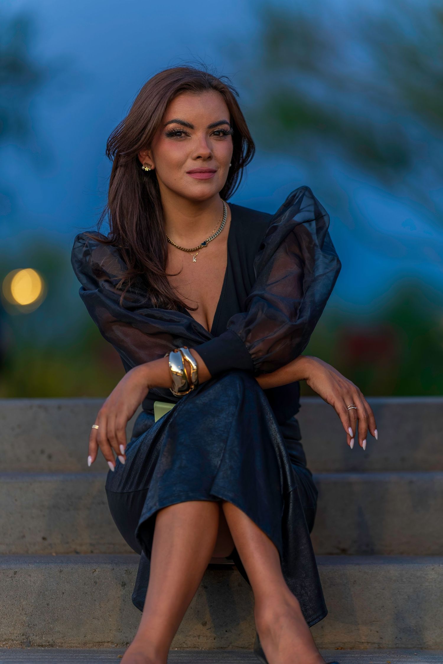 Elegant woman in black dress with gold jewelry sitting on stone steps at dusk.