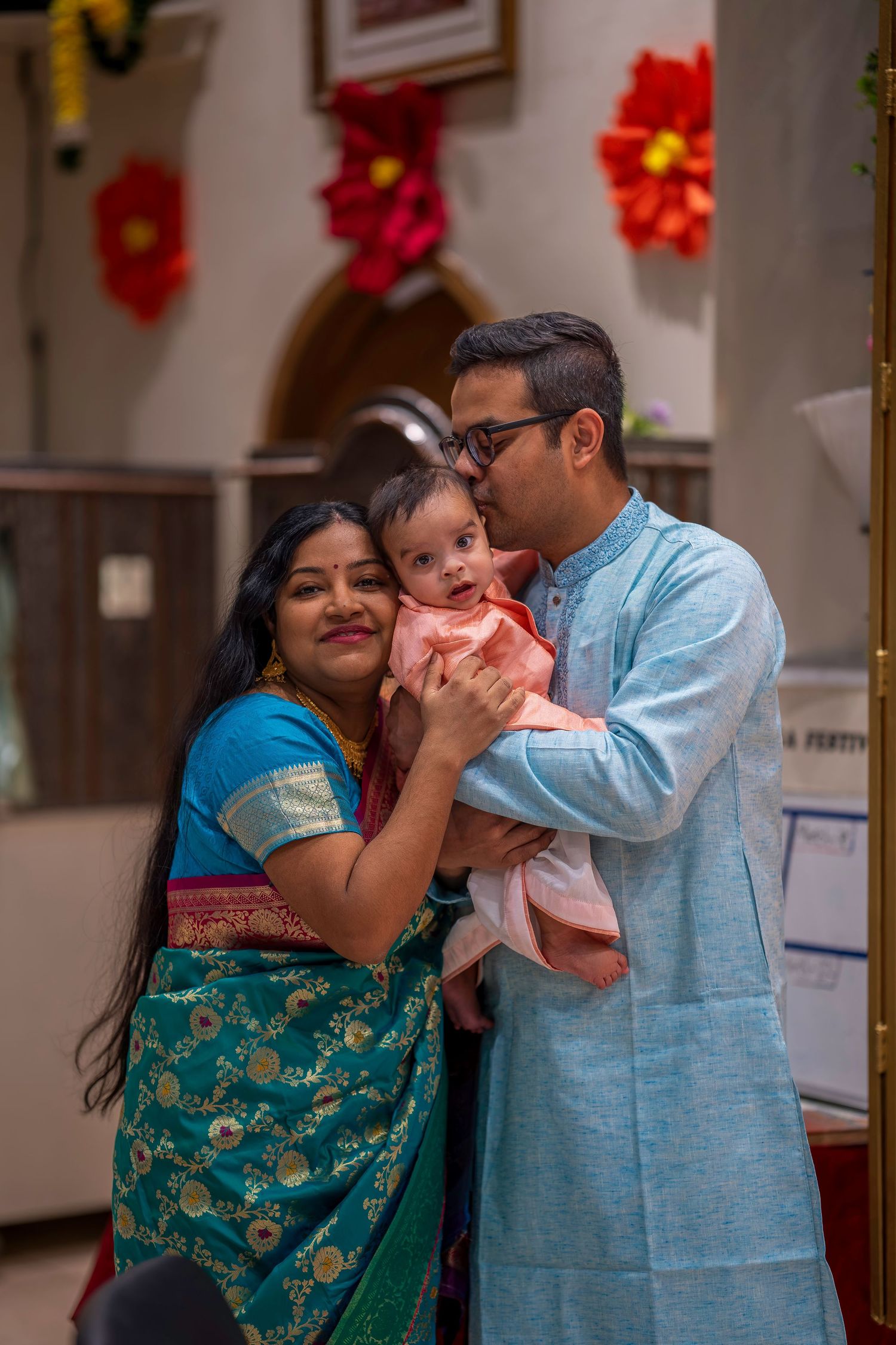Indian family in traditional attire, parents holding baby at festive celebration, man kissing infant's head.