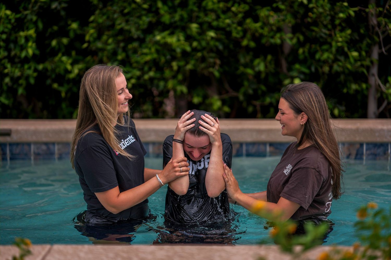 Two women assist a person during a baptism ceremony in an outdoor pool surrounded by lush greenery.