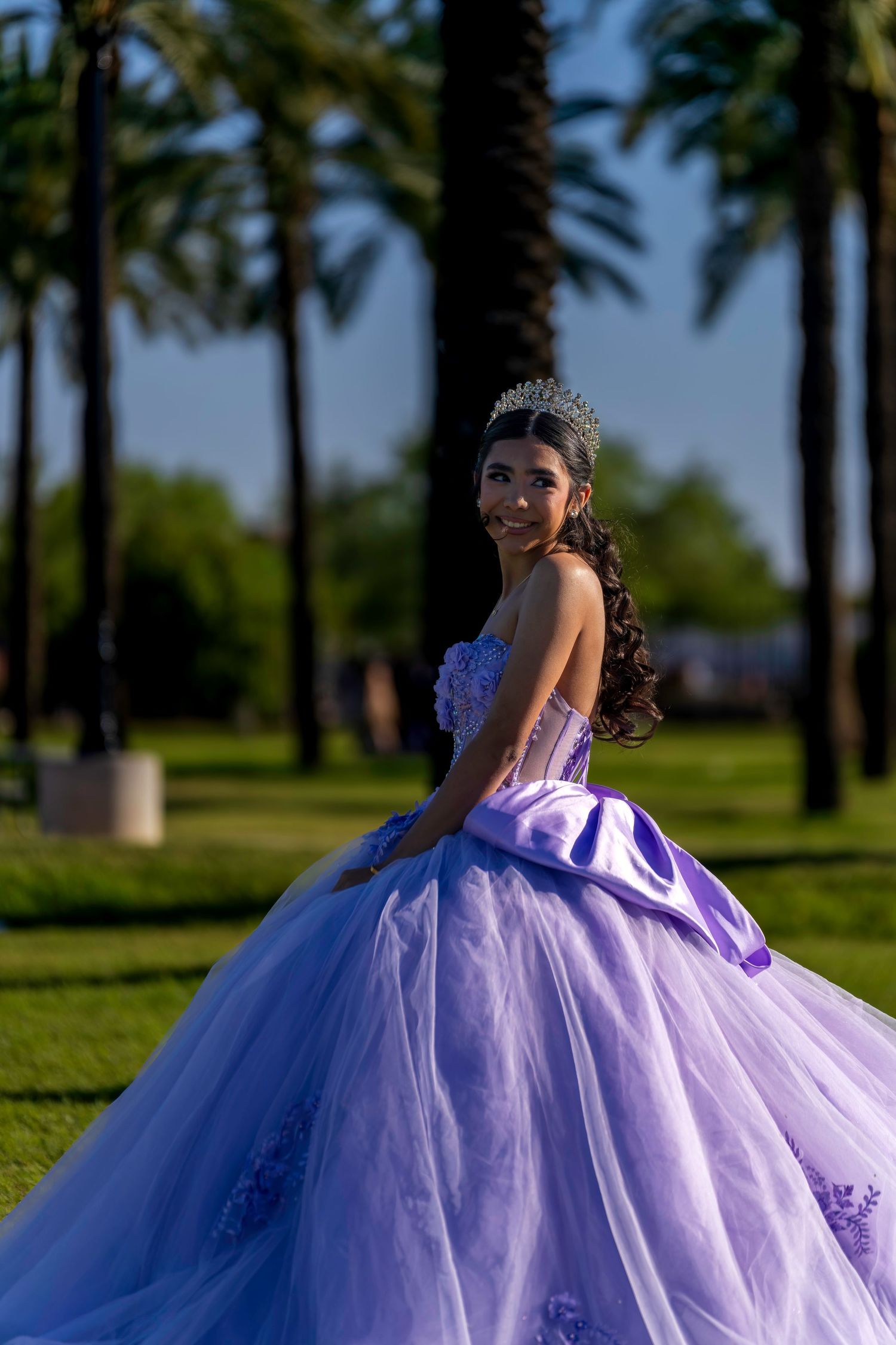 Young woman in lavender quinceañera ball gown and tiara poses gracefully outdoors among tall palm trees.