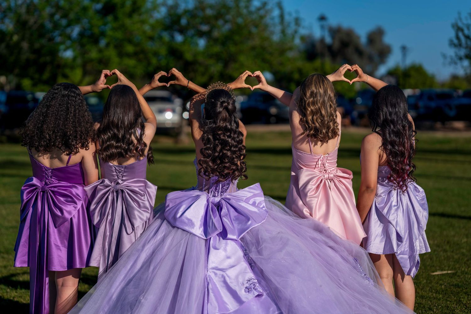 Five girls in purple and pink formal gowns with bows make heart shapes with their hands at an outdoor quinceañera.