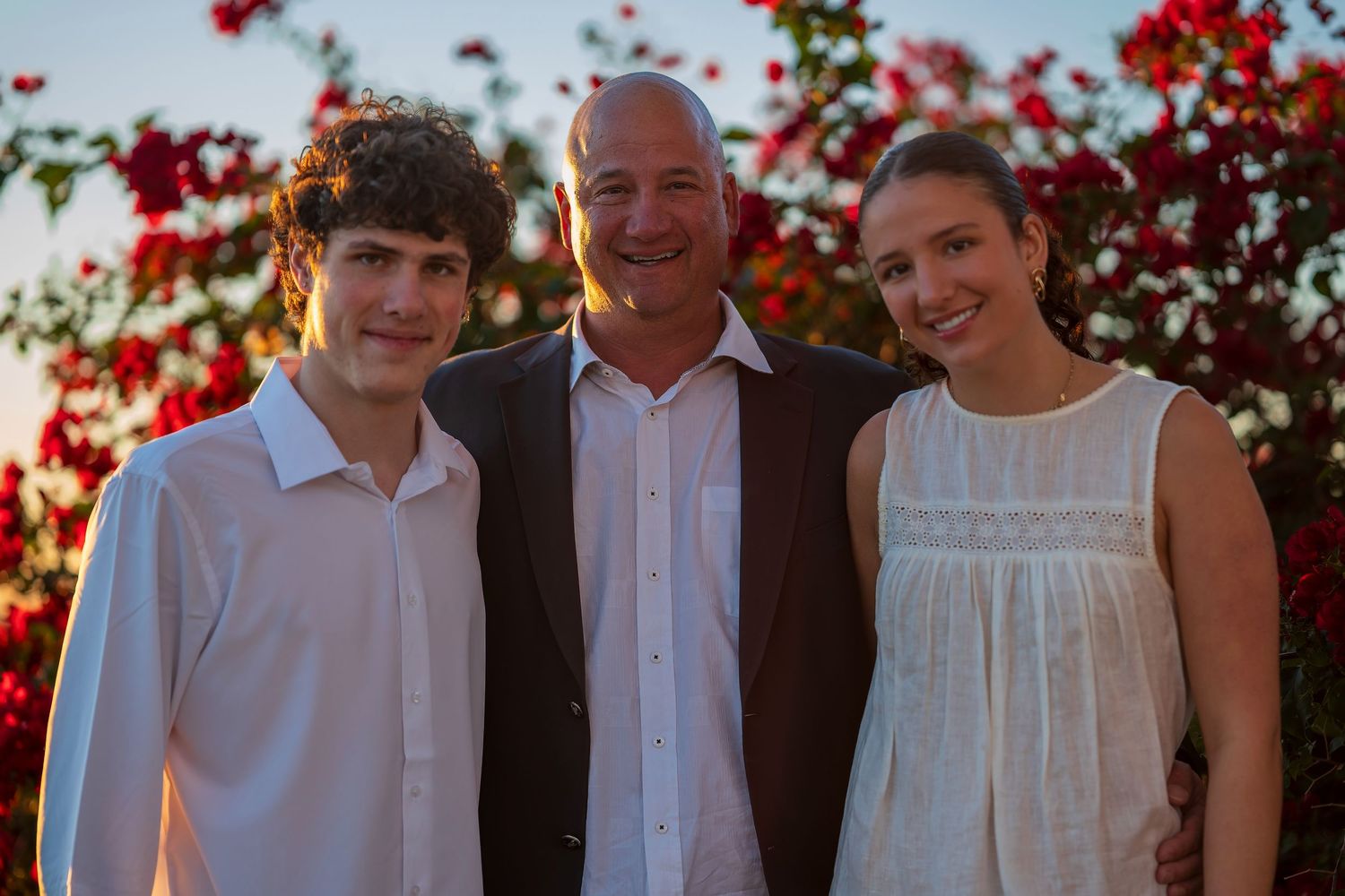 Family group photo with red flowering bush and evening sky in background.