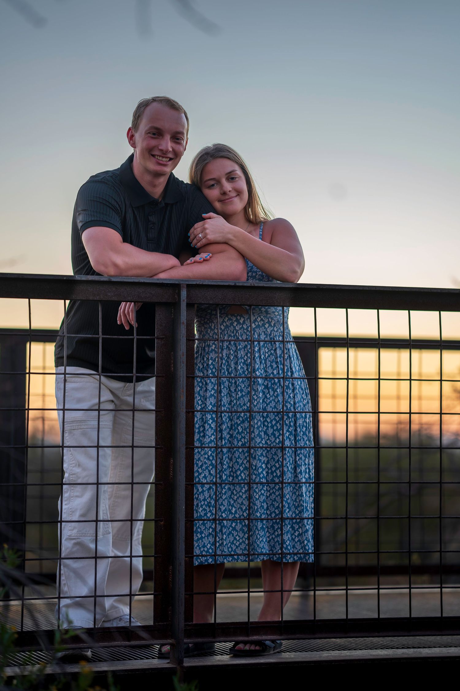 Smiling couple leaning on metal railing at sunset, woman in blue floral dress embracing man in black shirt.