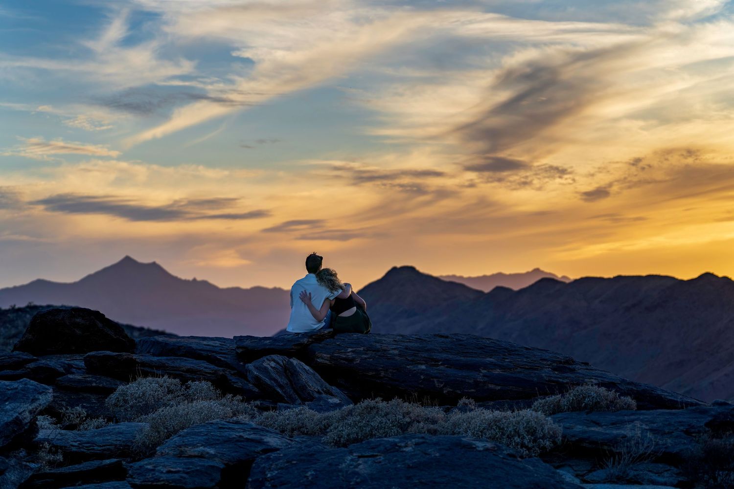 Two people sitting on rocks embracing while watching a breathtaking mountain sunset together.