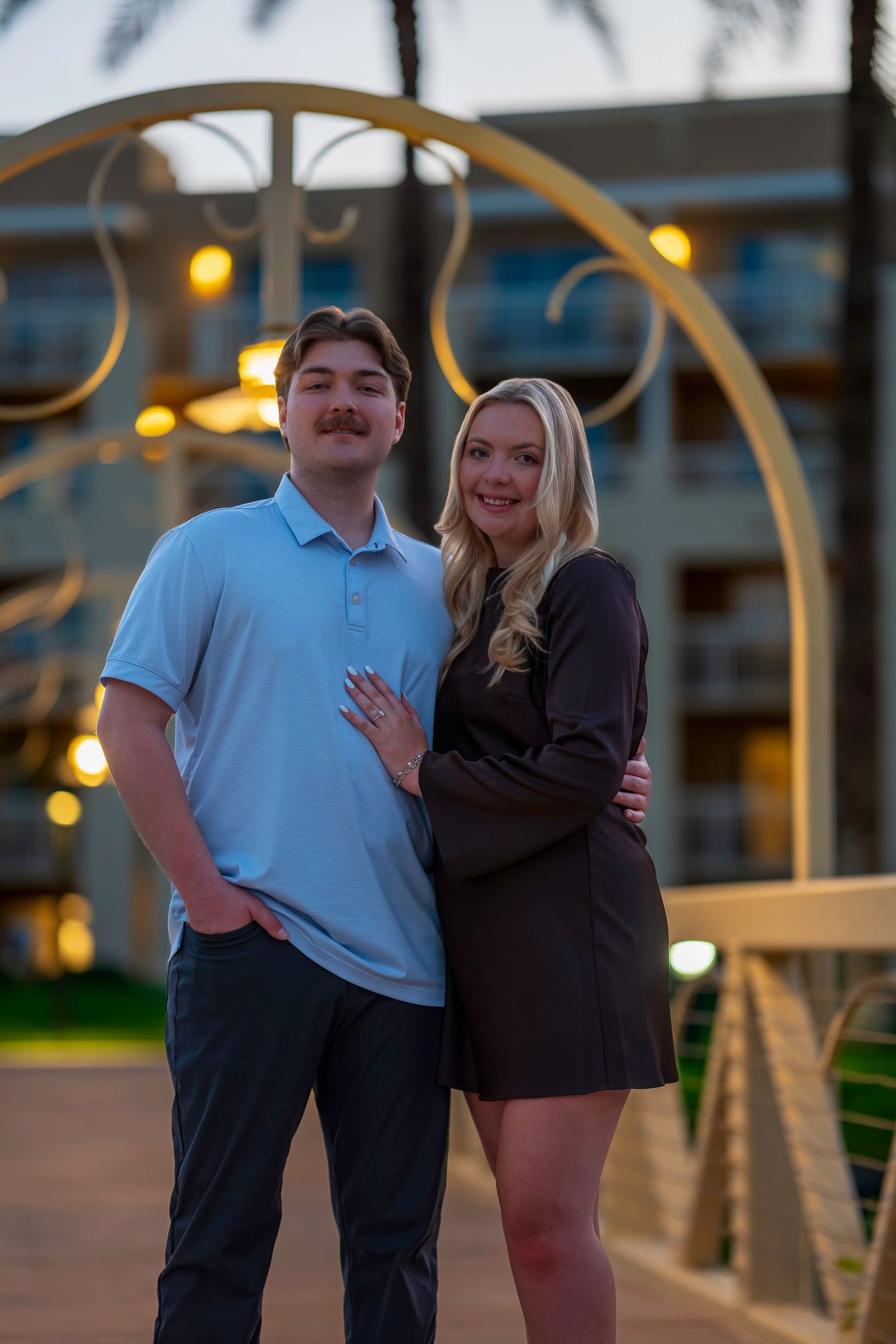 Couple posing together on a bridge at twilight, woman showing engagement ring near resort.