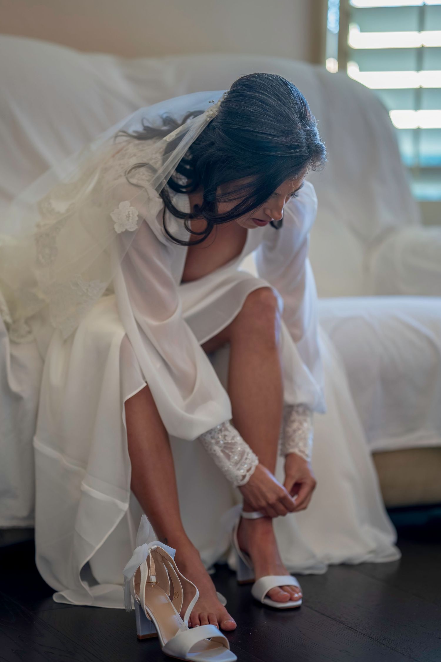 Bride in white gown and veil fastening her white heeled sandals while seated indoors before her wedding.