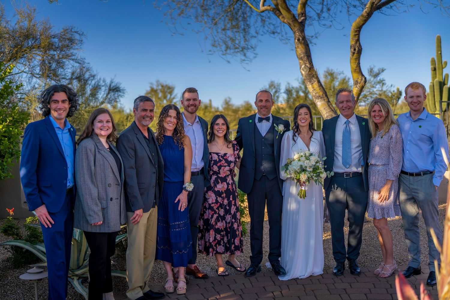 Bride and groom pose with family group of ten in desert garden setting at outdoor wedding.