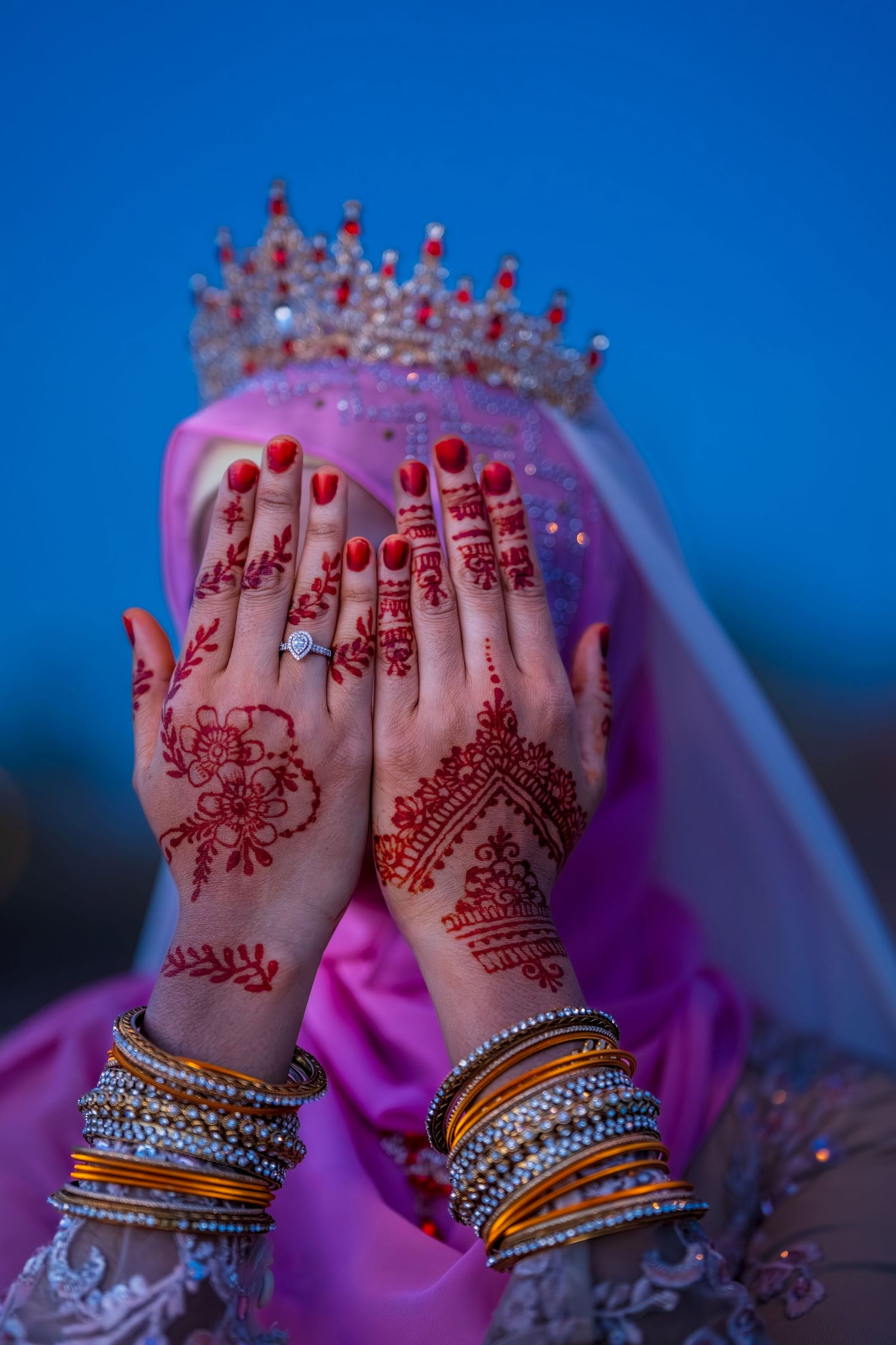 Muslim bride in pink hijab and crown displays intricate red henna mehndi designs on hands with gold bangles.