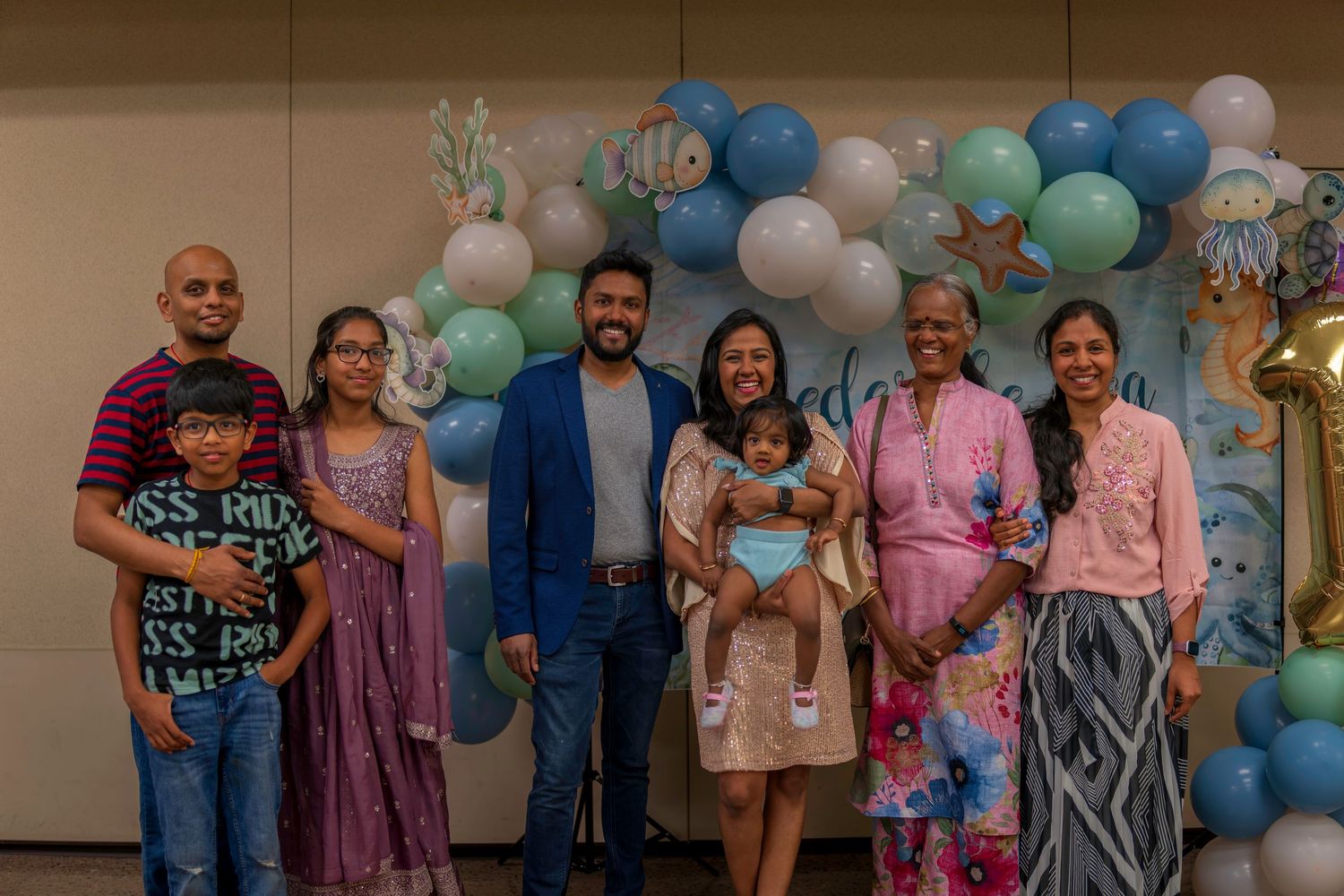 Family group photo at a first birthday party with balloon arch backdrop in blue, green, and white.